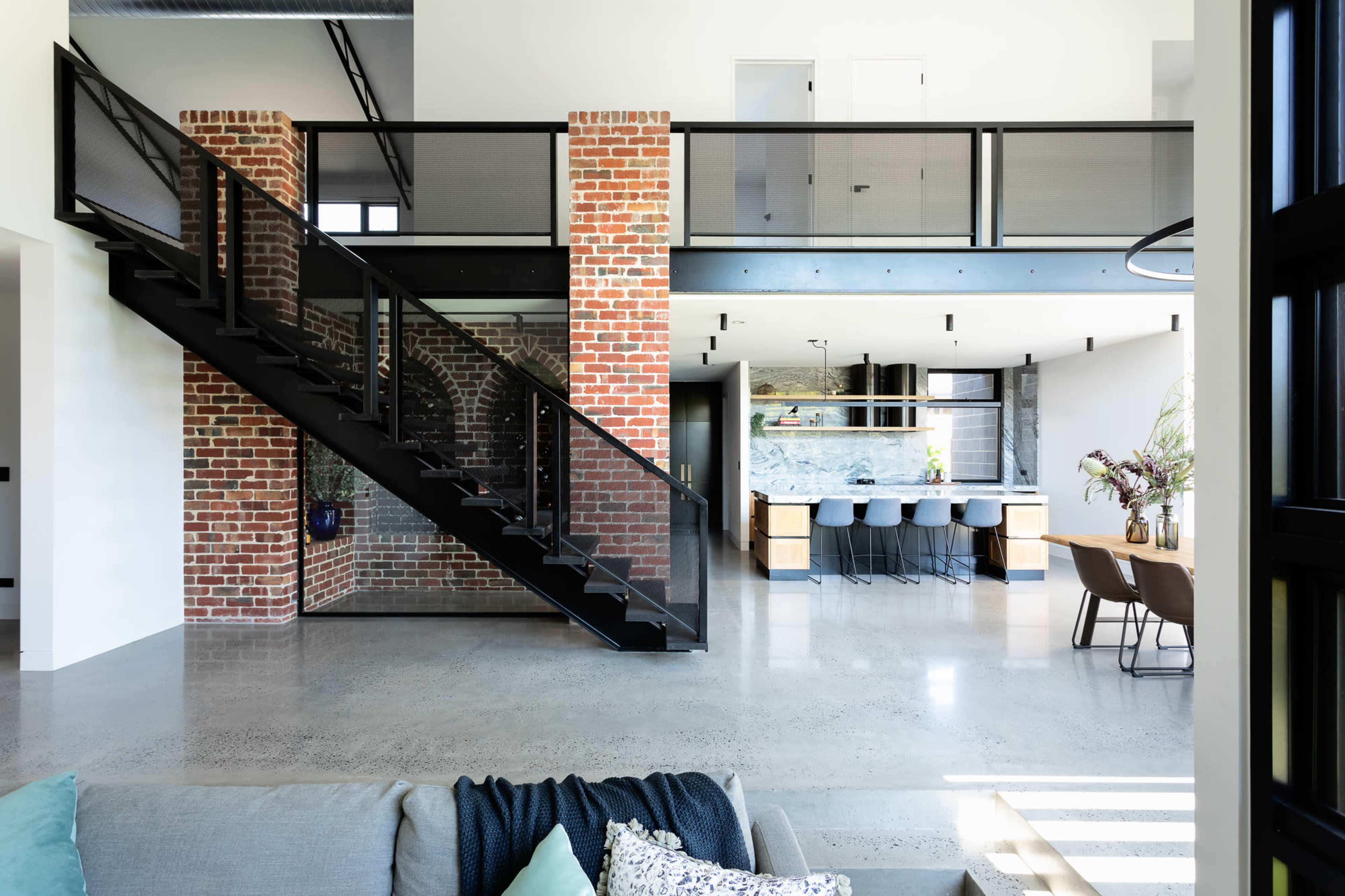 A modern interior with a black metal staircase leading to a mezzanine level, featuring exposed brick walls and an open kitchen and dining area with a large dining table and stools.