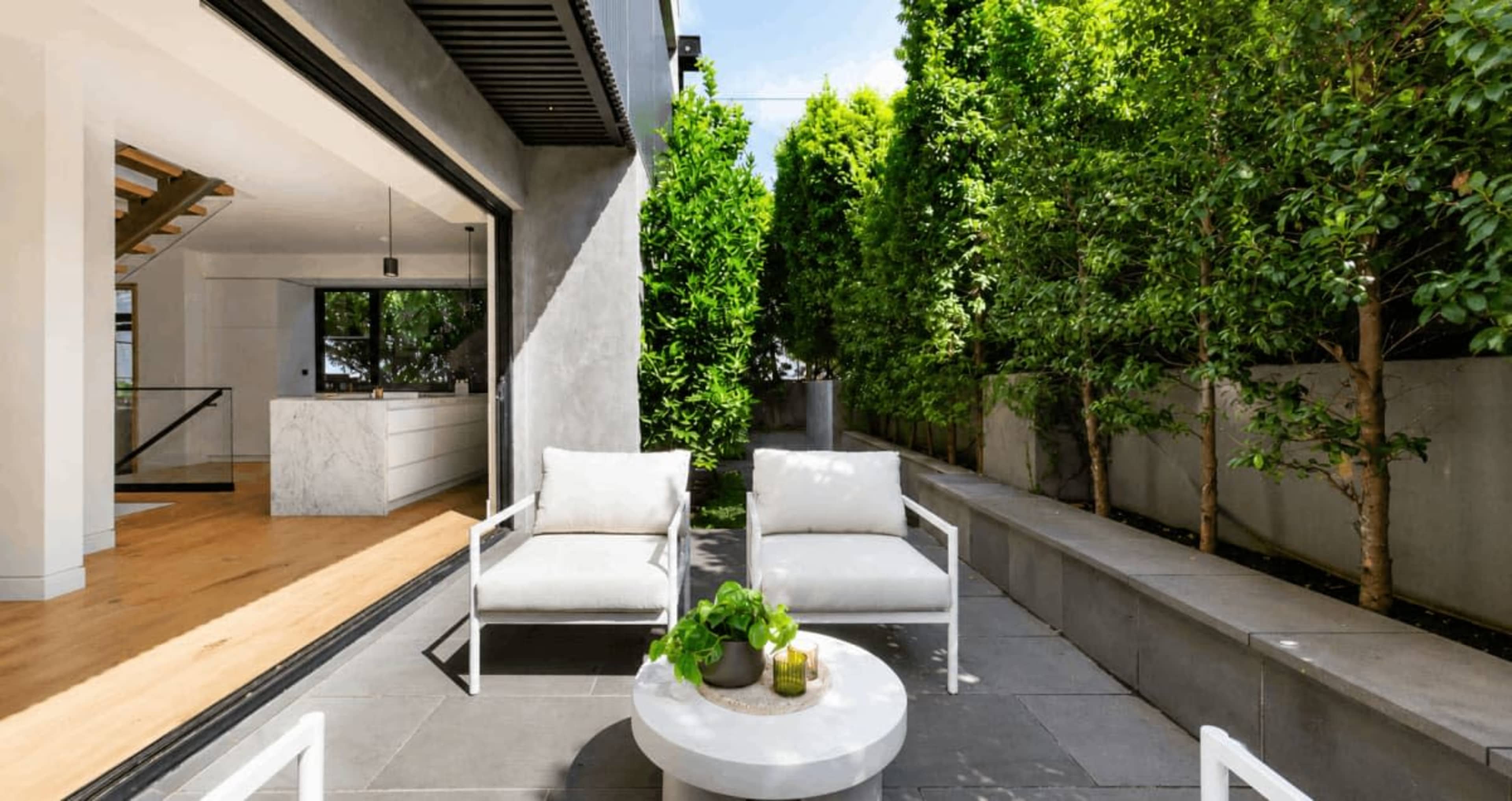 The image shows a modern outdoor seating area with two white chairs and a round table set against a backdrop of lush green hedges and an open doorway leading into a contemporary interior space.