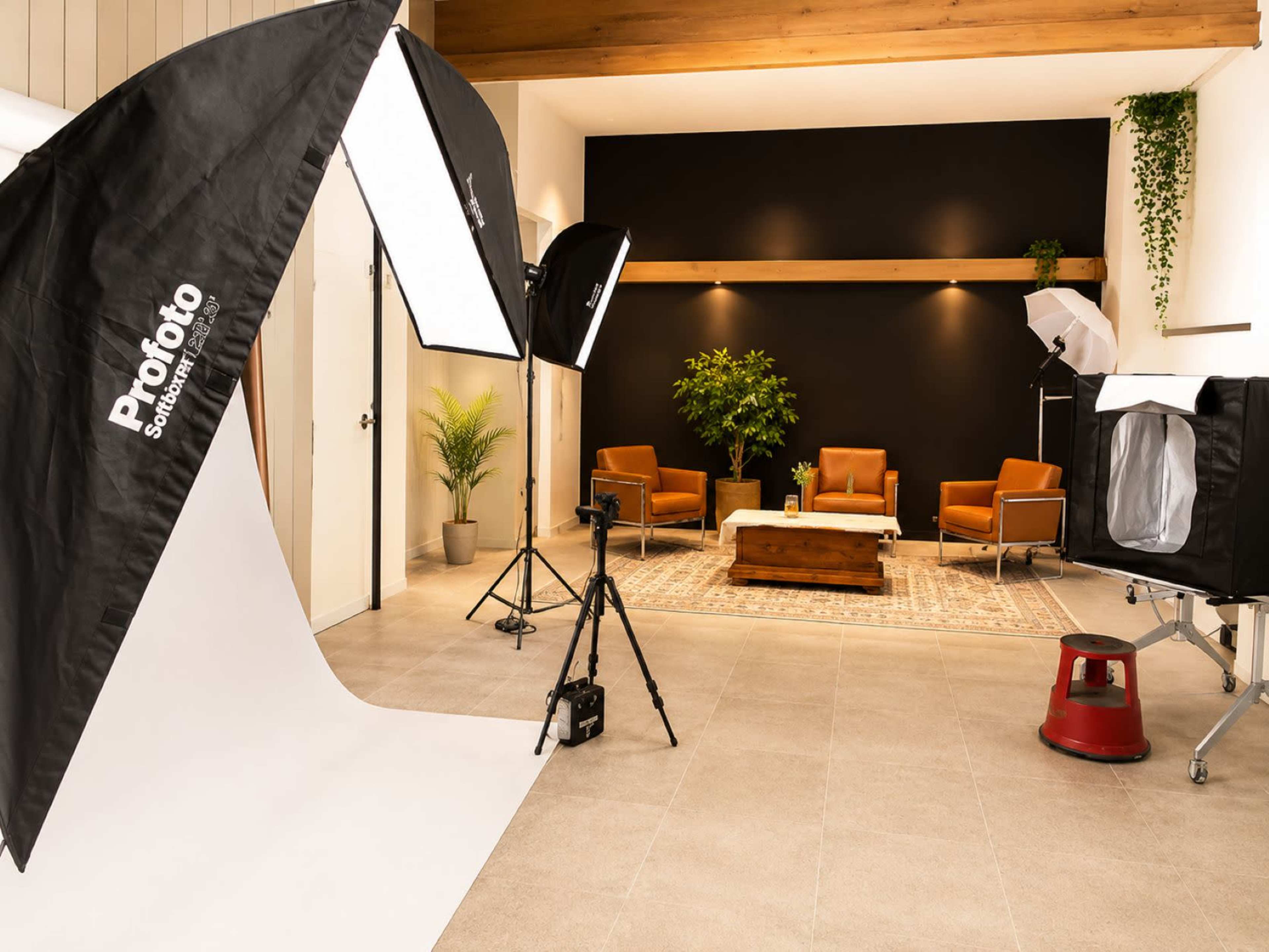The image shows a modern photo studio setup featuring two softbox lights, a backdrop, and a seating area with two brown chairs and a wooden coffee table.