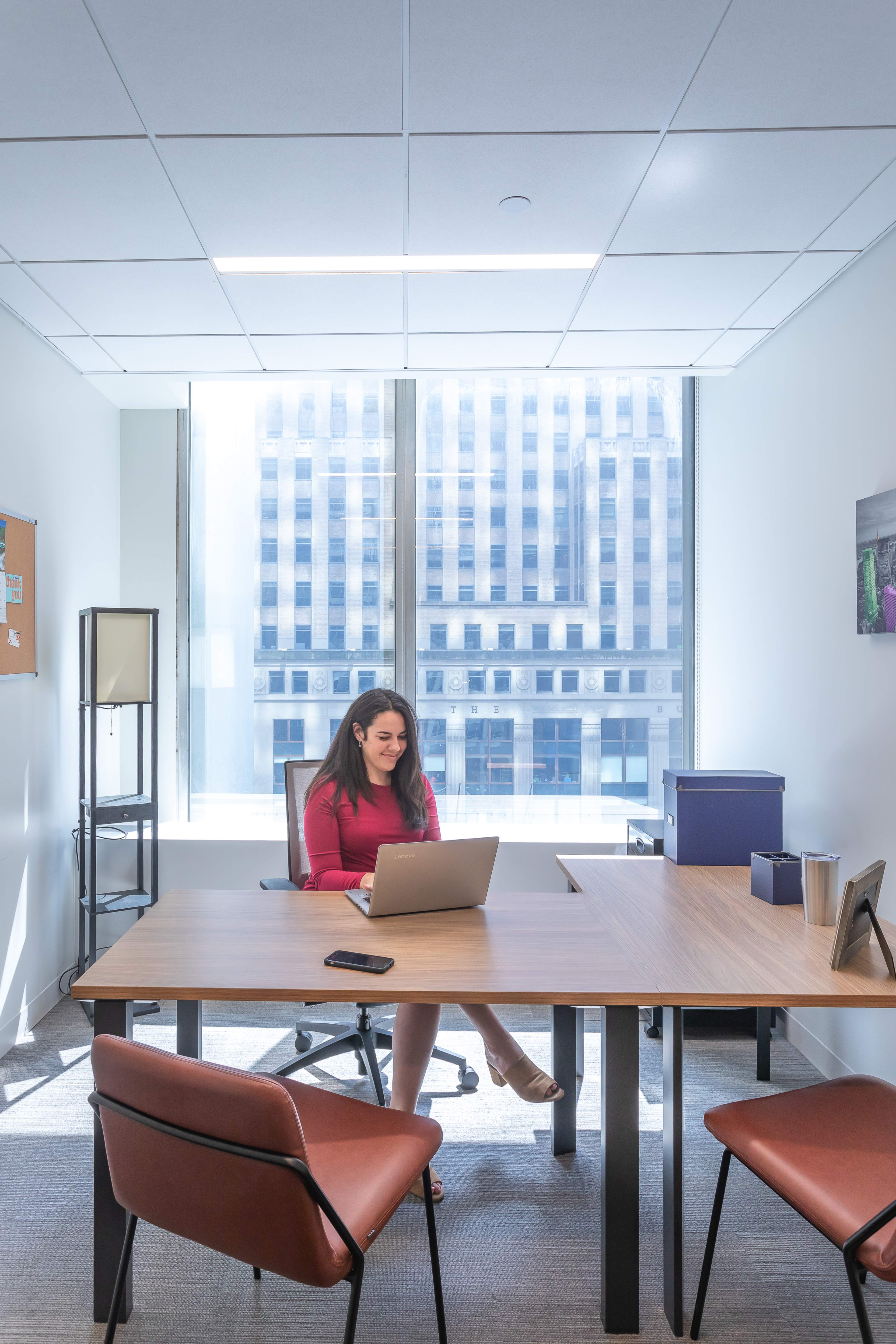 A woman in a red dress is sitting at a wooden desk with a laptop in a well-lit office with large windows.