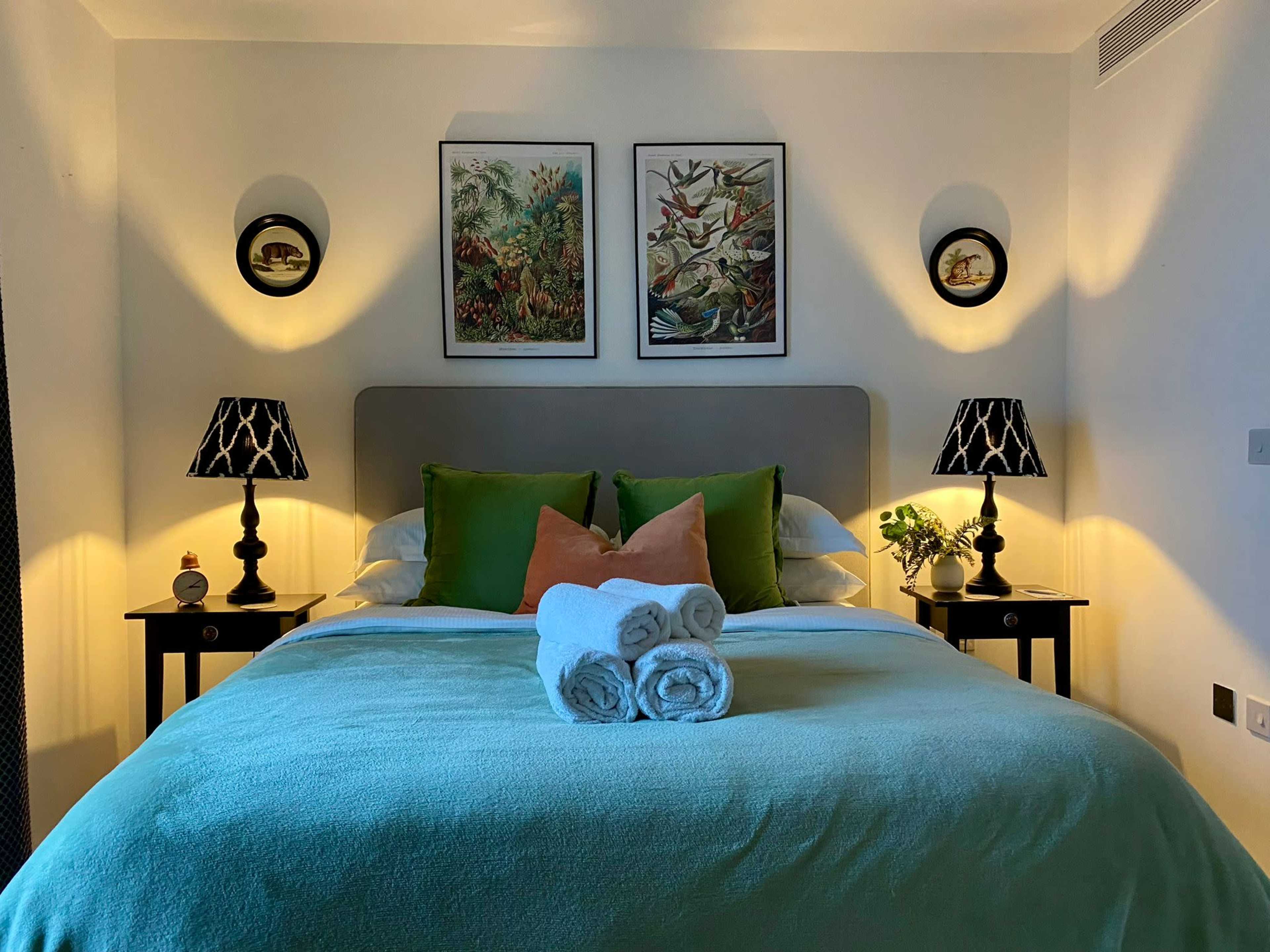 The image shows a well-decorated bedroom featuring a double bed with green and white bedding, two bedside tables with lamps, and artwork on the walls.