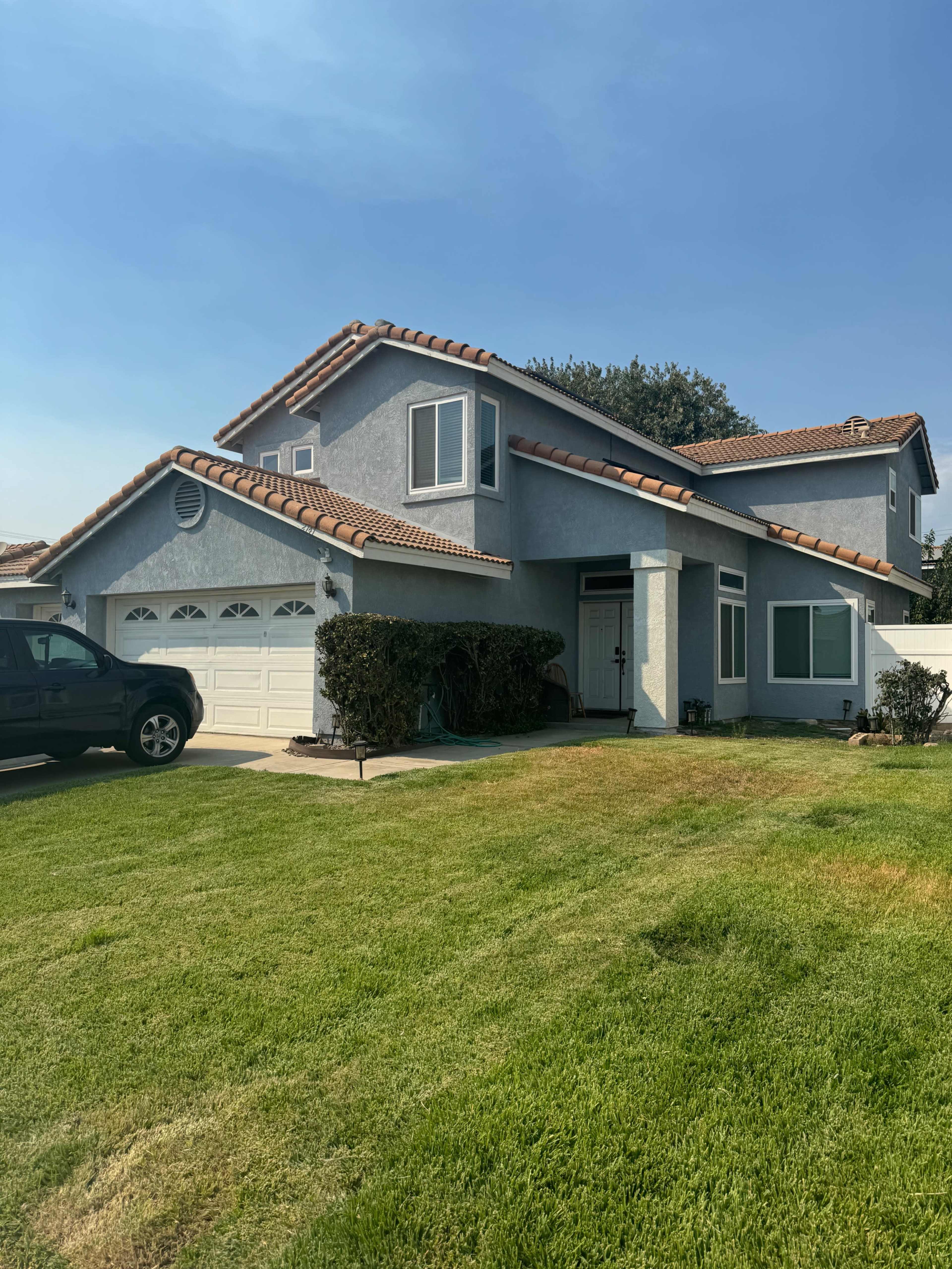 A two-story gray house with a red-tiled roof, a front lawn, and a black truck parked in the driveway.