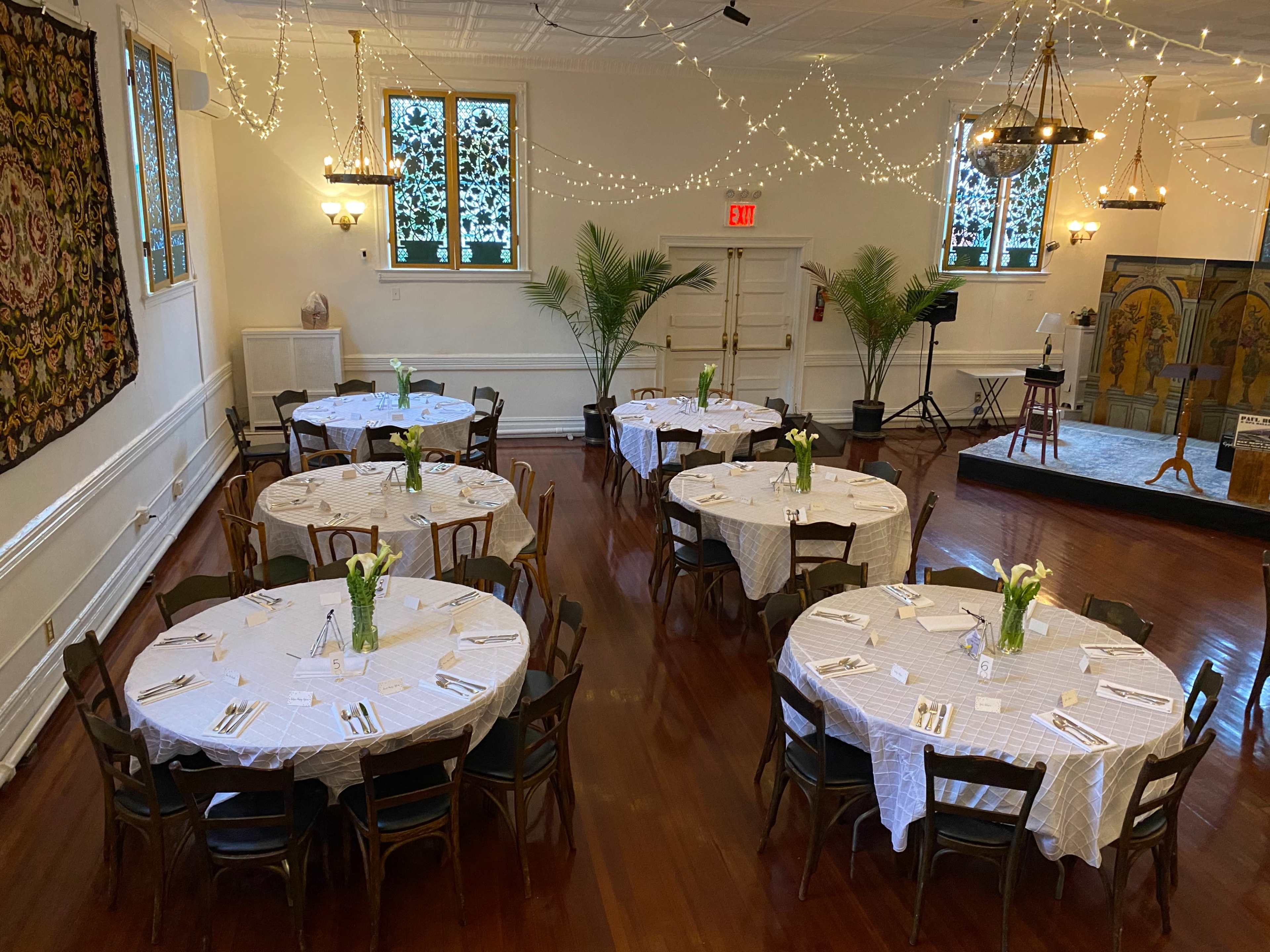 The image shows a large room set up for an event with several round tables covered with white tablecloths, arranged with utensils and floral centerpieces, under soft lighting and decorative string lights.