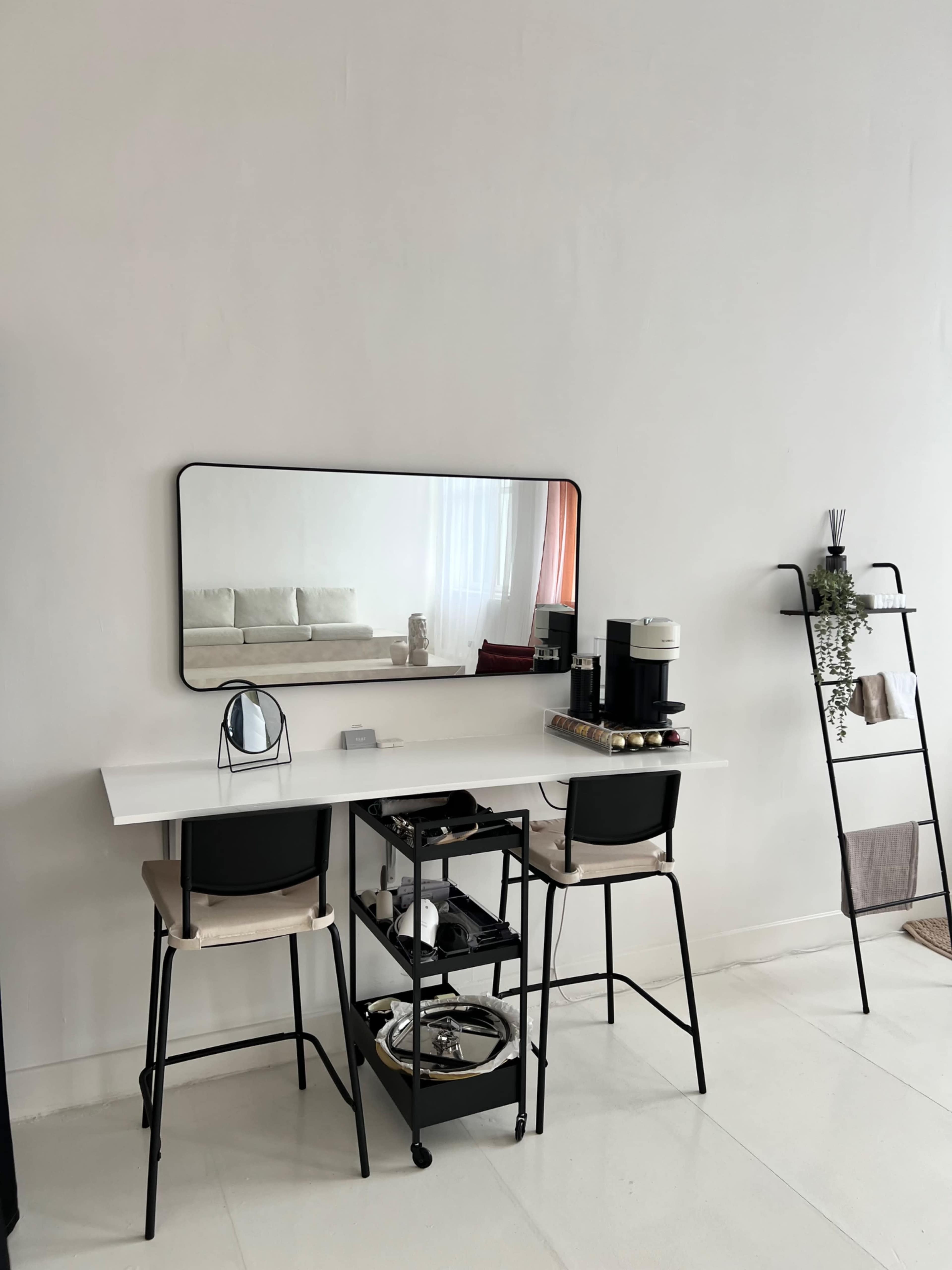 The image shows a minimalist dining area featuring a white table with two black stools, a large rectangular mirror on the wall, and a mobile cart with kitchen items beside a window.