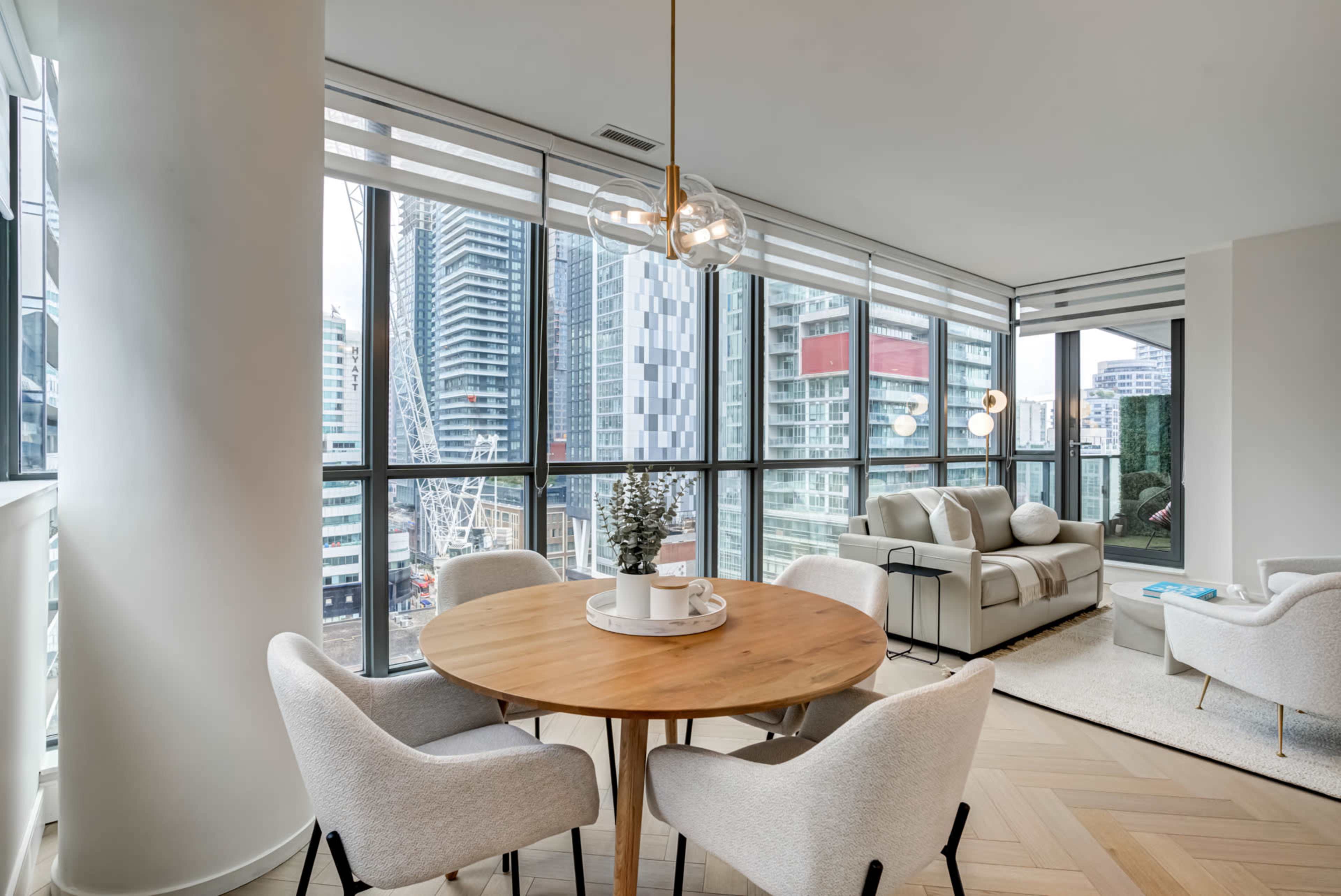 A dining area features a round wooden table surrounded by four upholstered chairs, with large windows showcasing a cityscape and modern furniture in the living space.