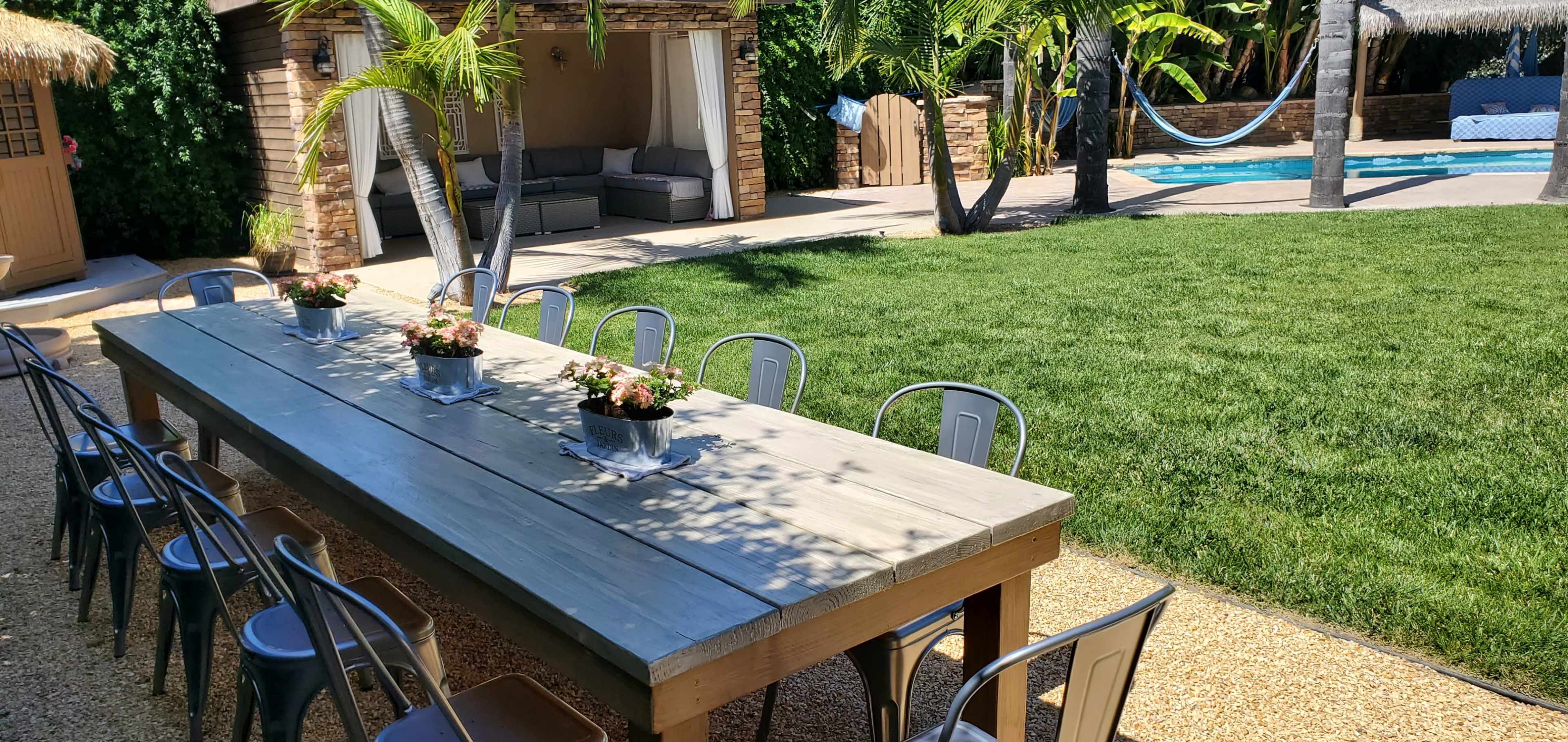 A large wooden dining table with metal chairs is set in a landscaped yard featuring a pool, hammock, and tropical plants.