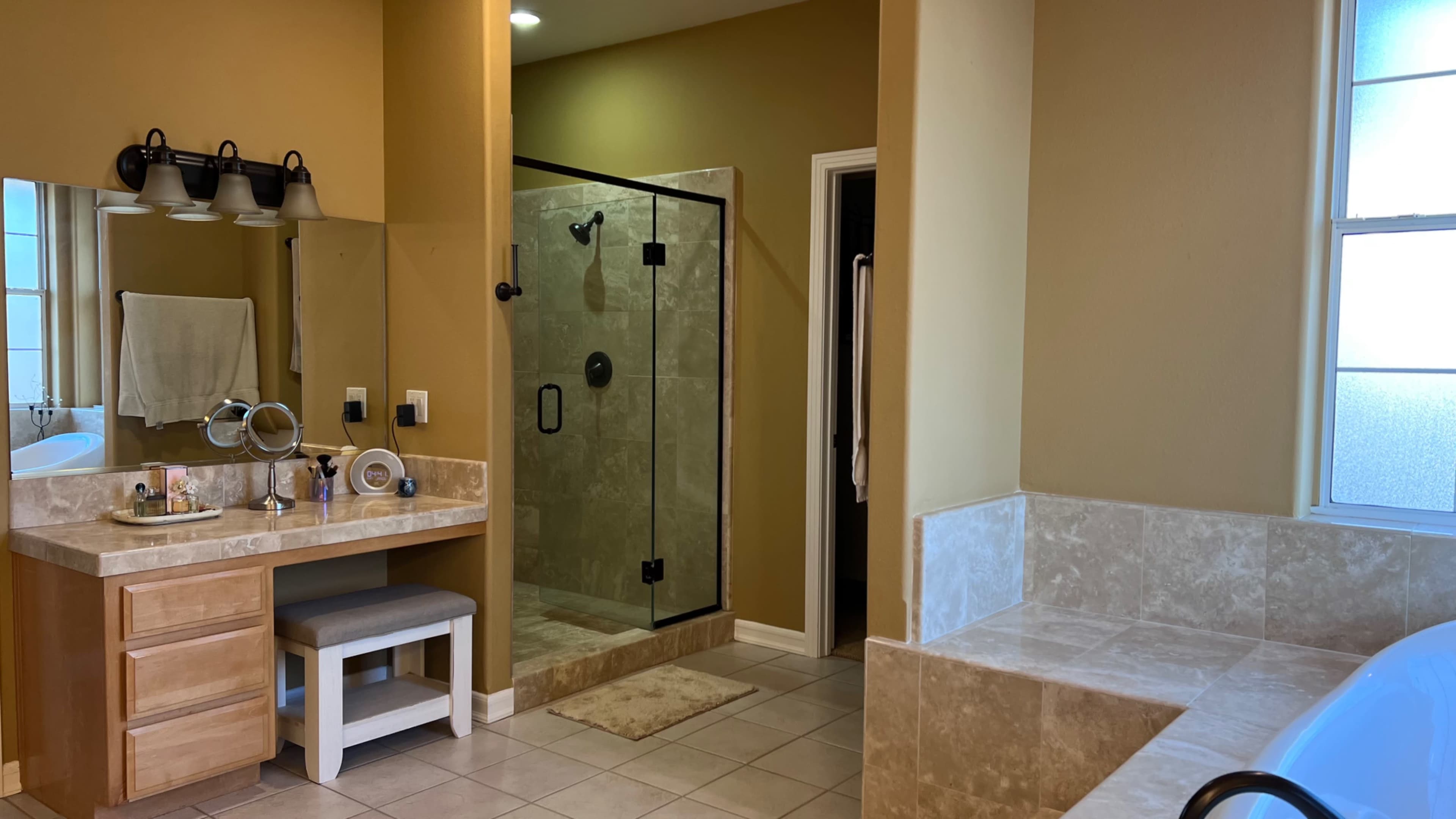 A spacious bathroom featuring a glass shower enclosure, a freestanding bathtub, a vanity with a mirror, and beige tile flooring.