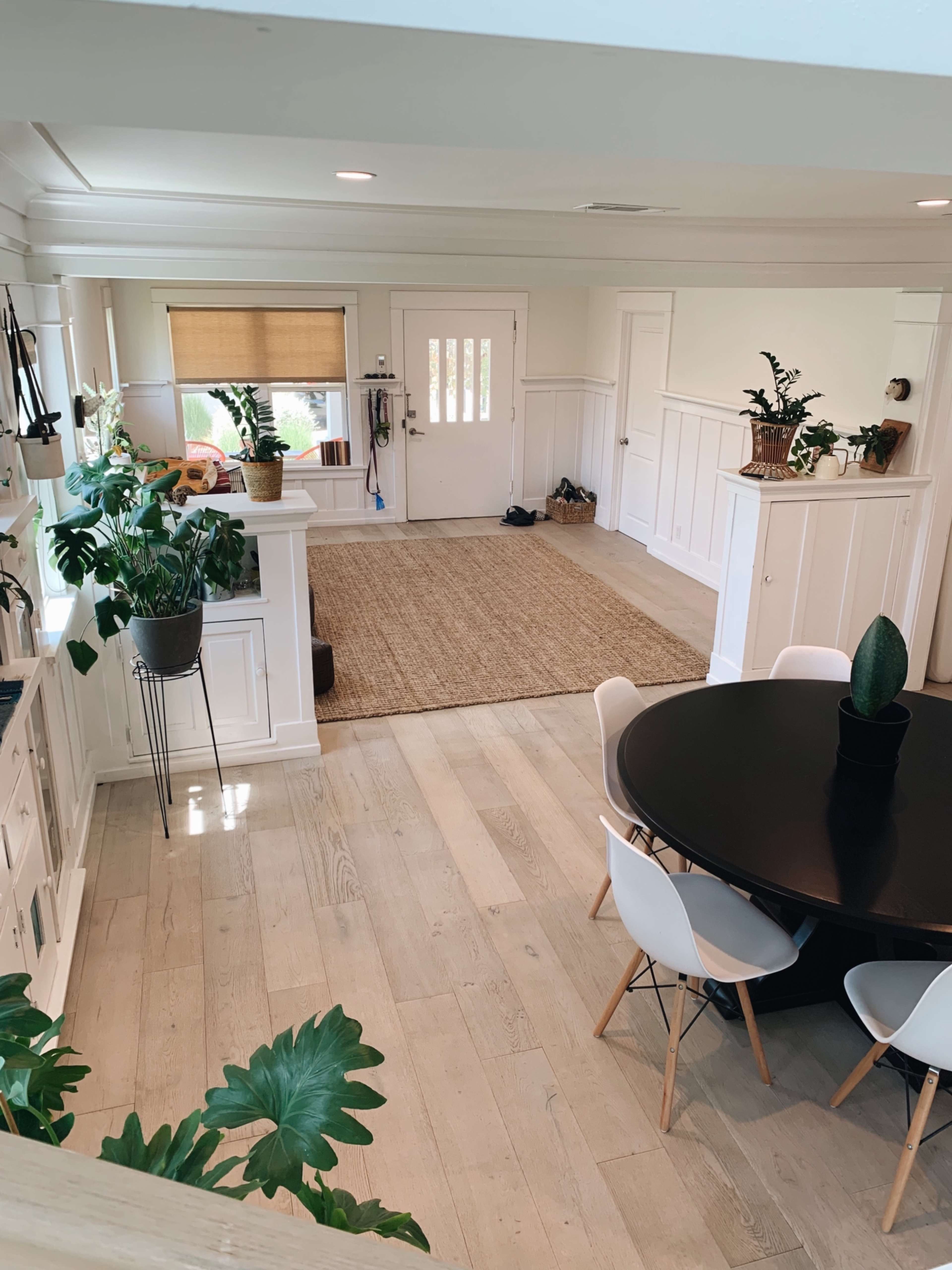 A bright, open interior space featuring a dining area with a black circular table and white chairs, along with a small entryway and several potted plants.