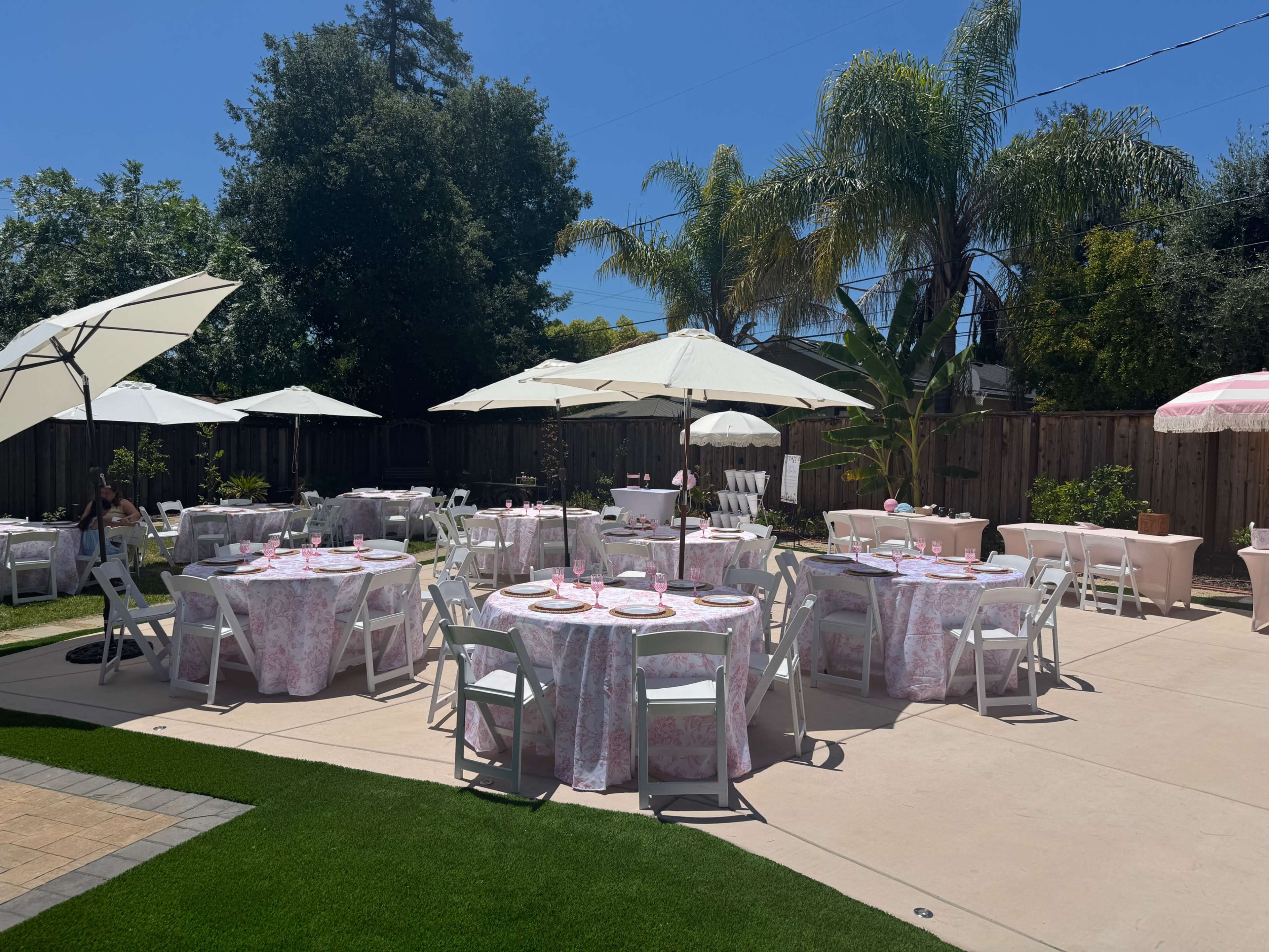 The scene features multiple round tables covered with pink floral tablecloths, accompanied by white chairs and large umbrellas, set up in a backyard area surrounded by greenery and blue skies.