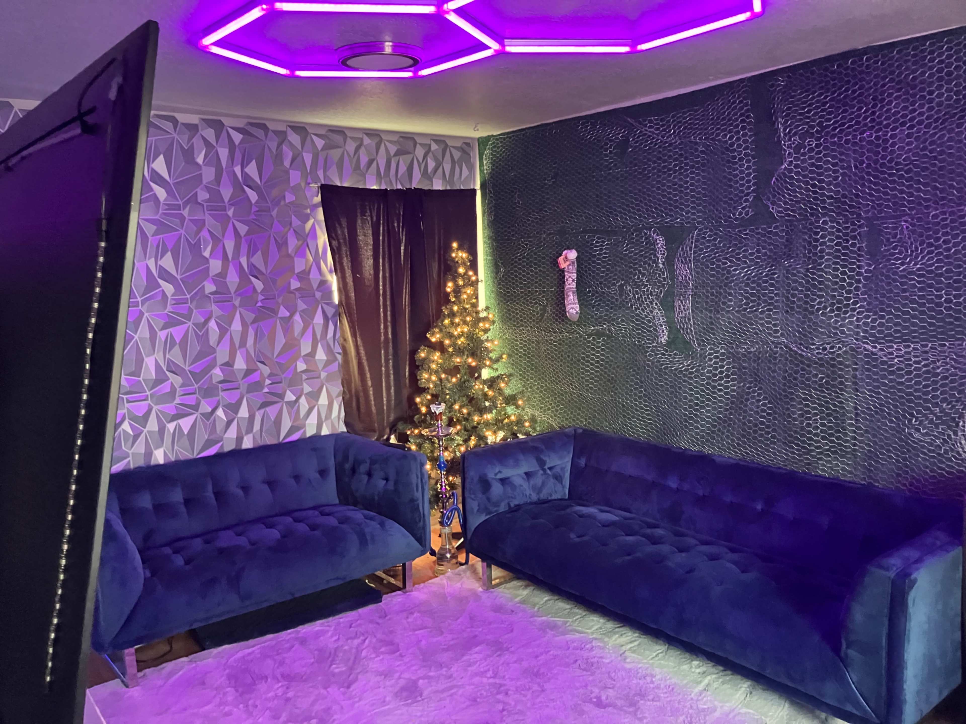 The scene features a modern living room with two blue velvet sofas, a decorated Christmas tree, and a wall adorned with patterned wallpaper and purple LED lighting.