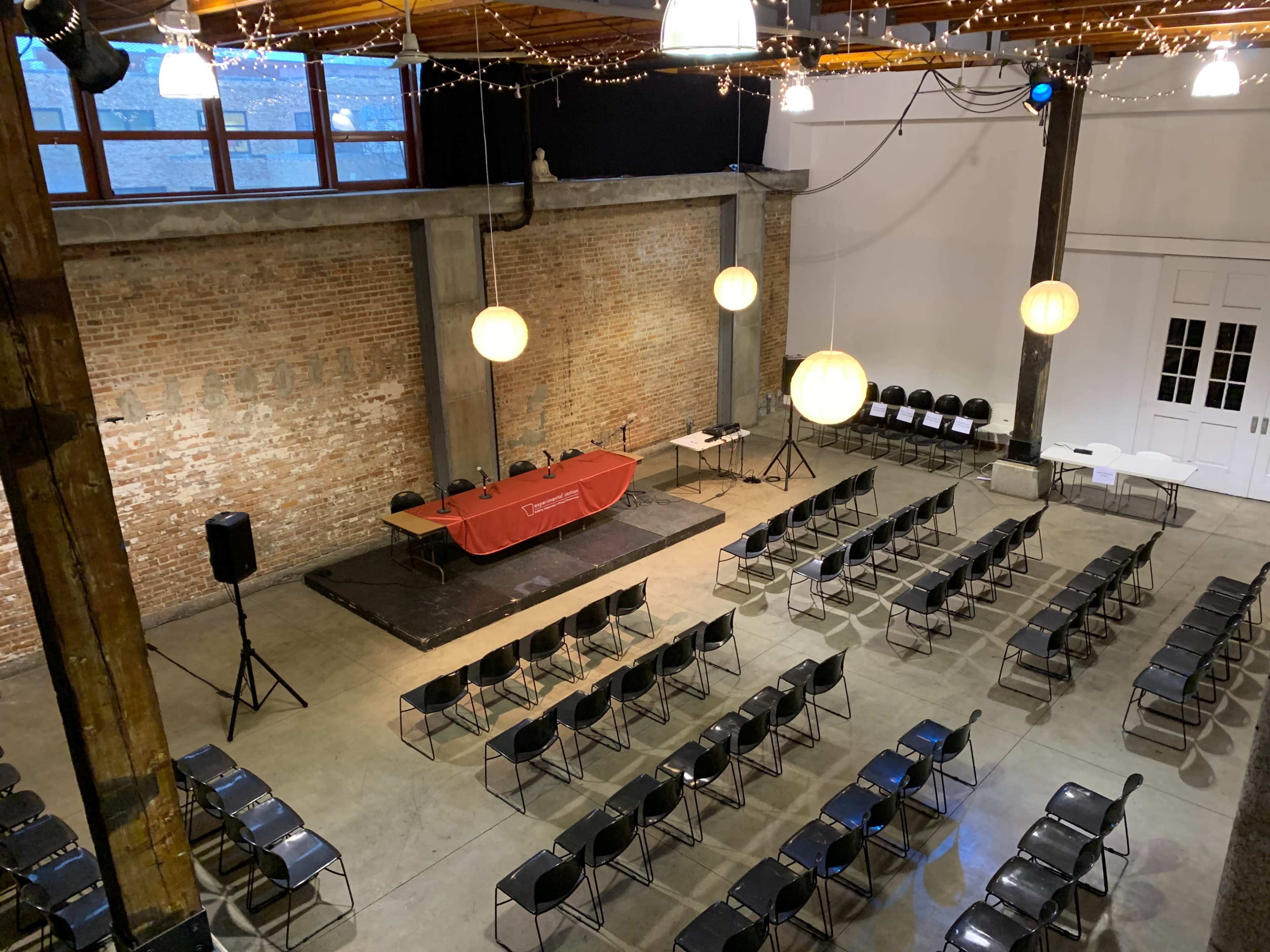 An empty event space with rows of black chairs facing a red table set up on a stage, accompanied by hanging light fixtures and exposed brick walls.