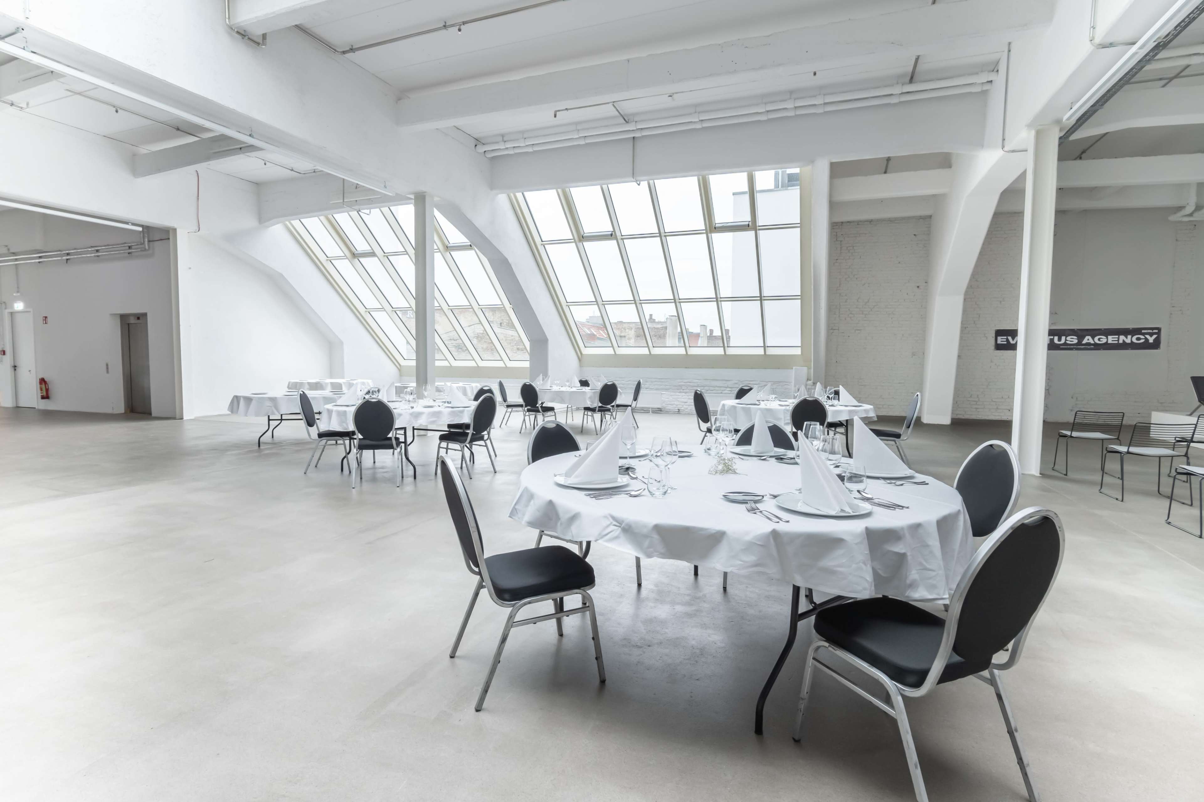 A spacious, modern event space features neatly arranged round tables with white tablecloths and chairs, illuminated by large windows.