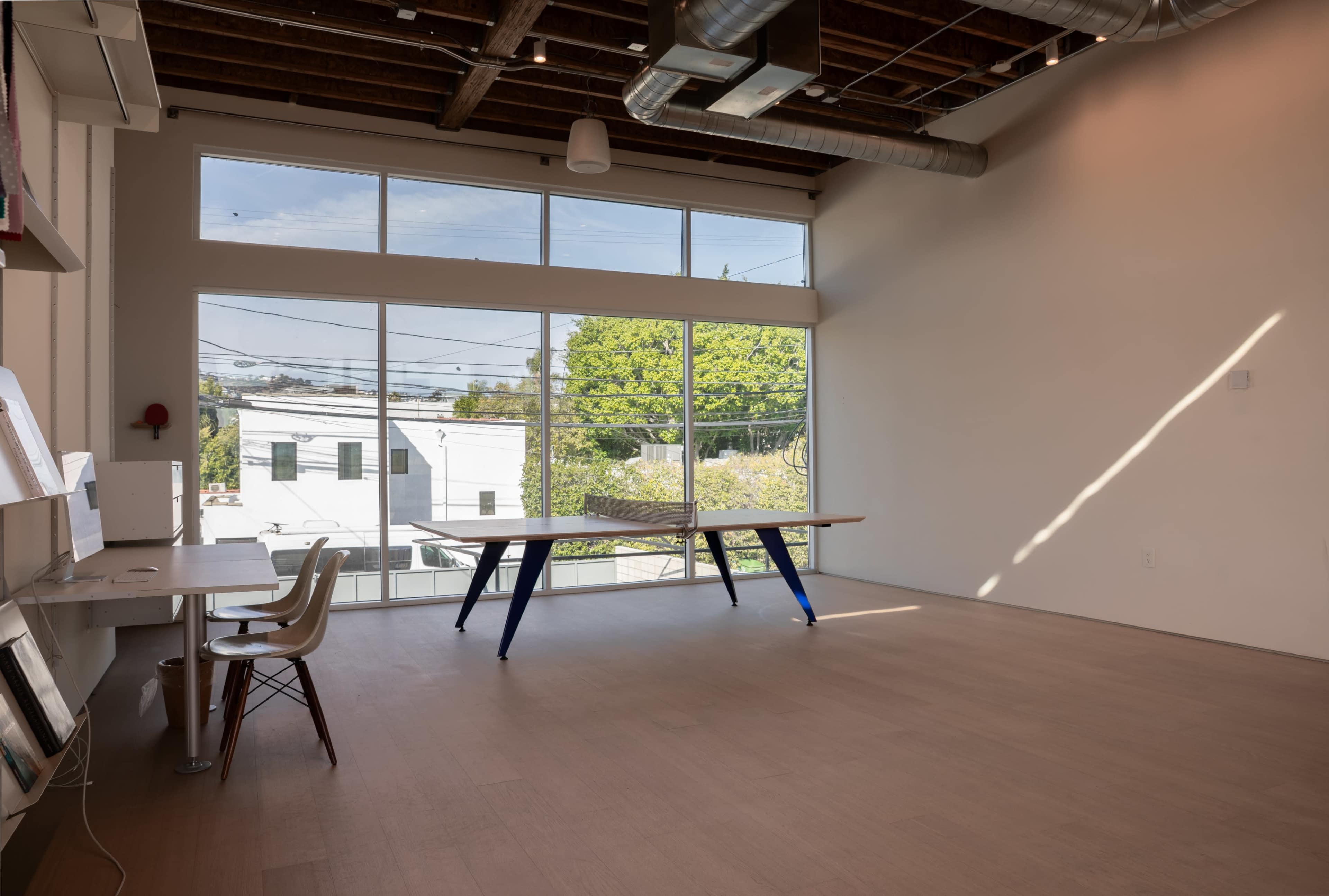 The image shows a bright, empty room with large windows, a central table, and minimal furnishings.