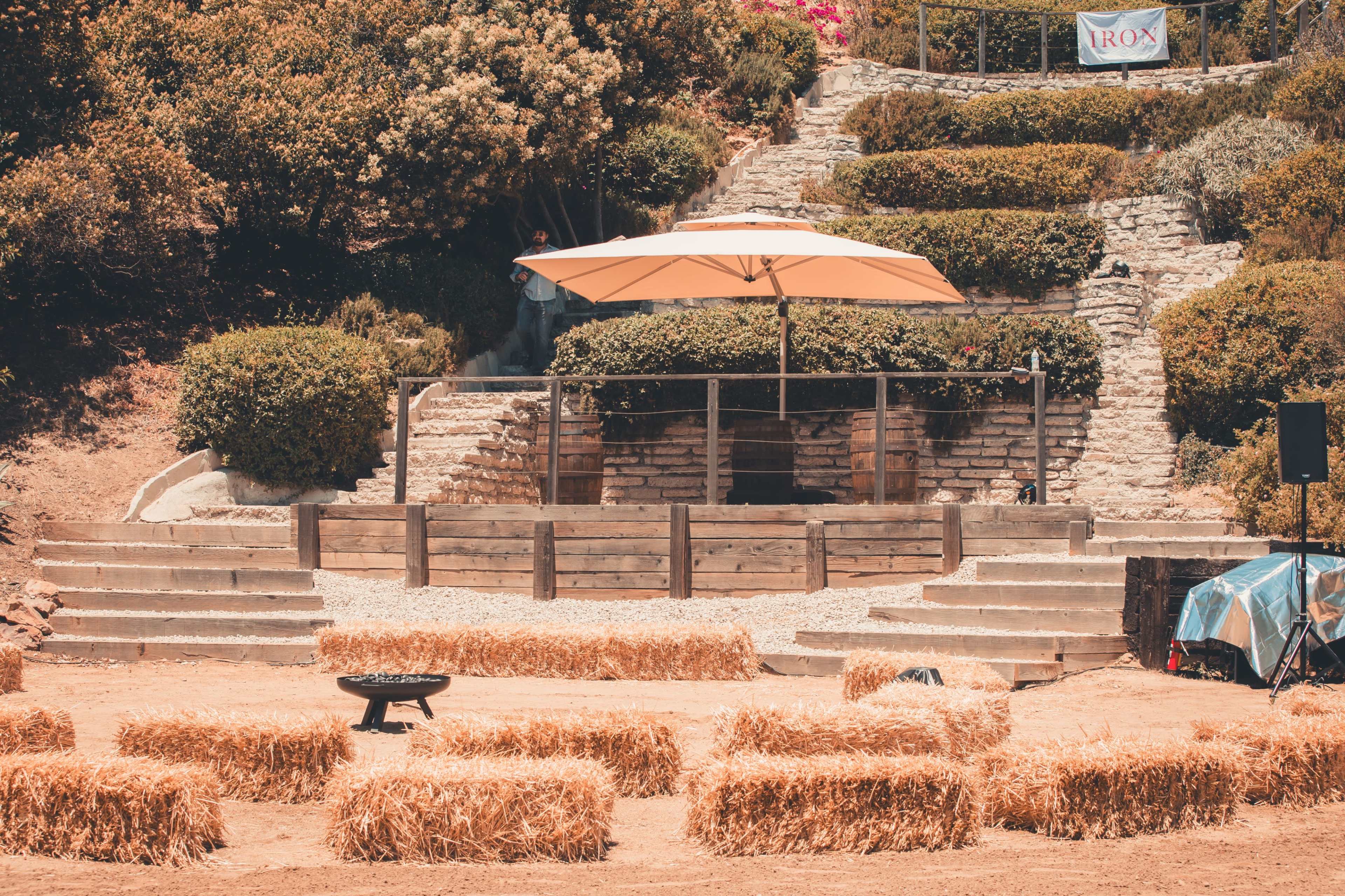 The image shows a rustic outdoor stage area with an umbrella, surrounded by bales of hay and landscaped greenery.