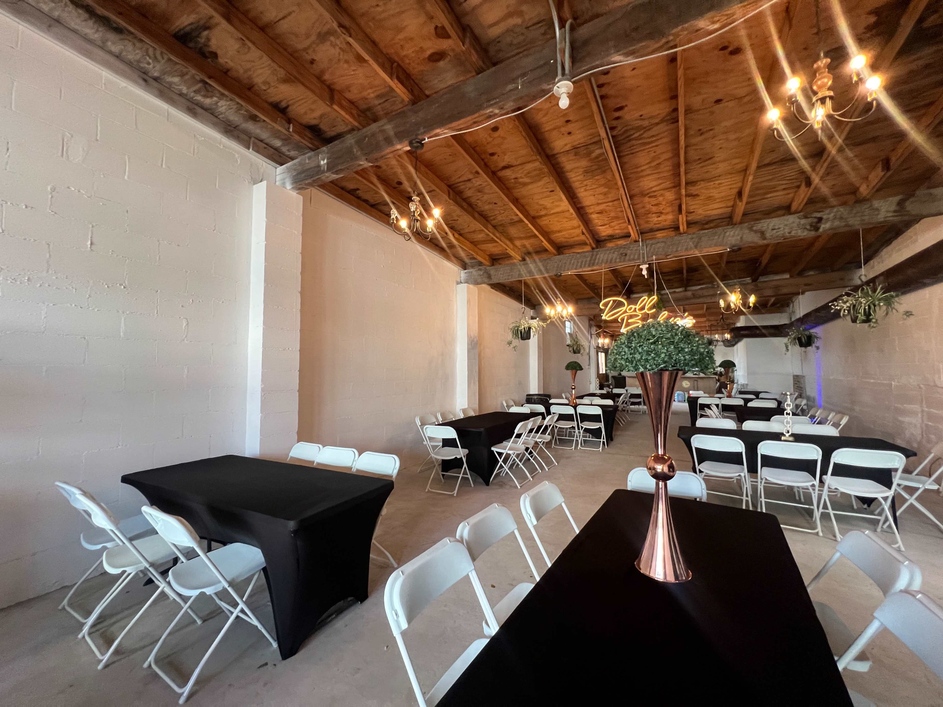 The image shows an indoor event space with white walls, wooden beams, and tables set with black tablecloths and white chairs.