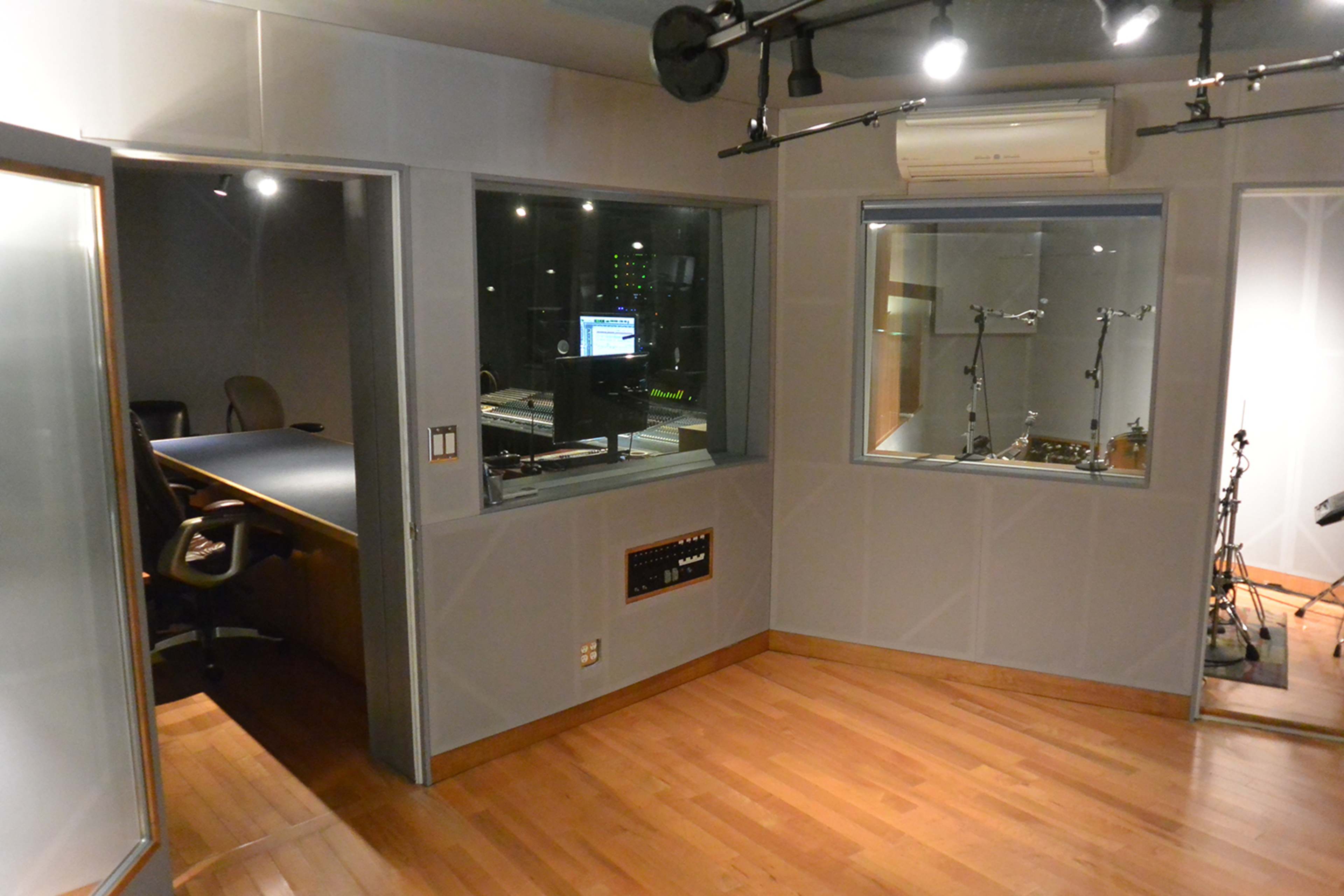 A music production studio with two separate soundproof rooms, one containing a desk and computer, and the other featuring microphone stands and instruments.