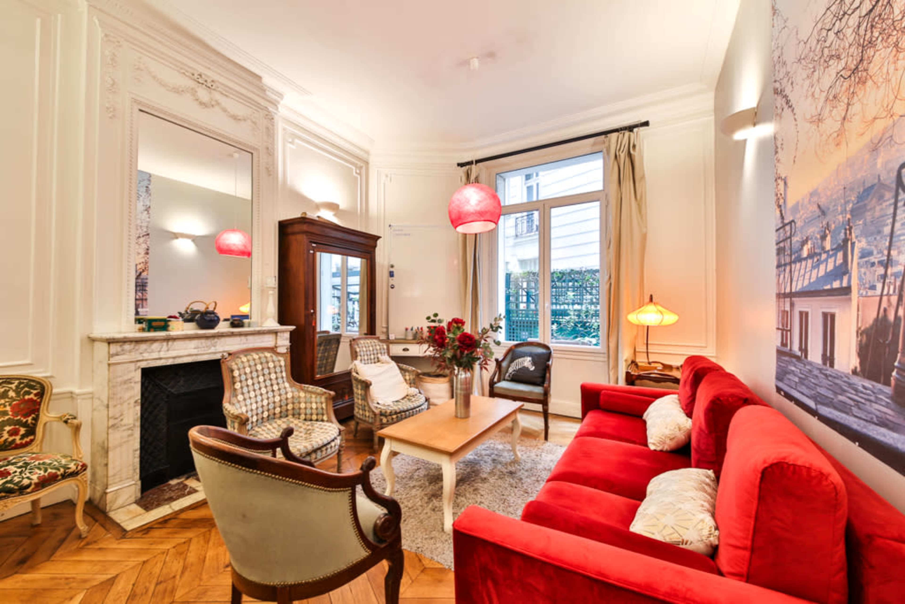 The image shows a cozy living room featuring a red sofa, patterned armchairs, a wooden coffee table, and a large window allowing natural light to enter.