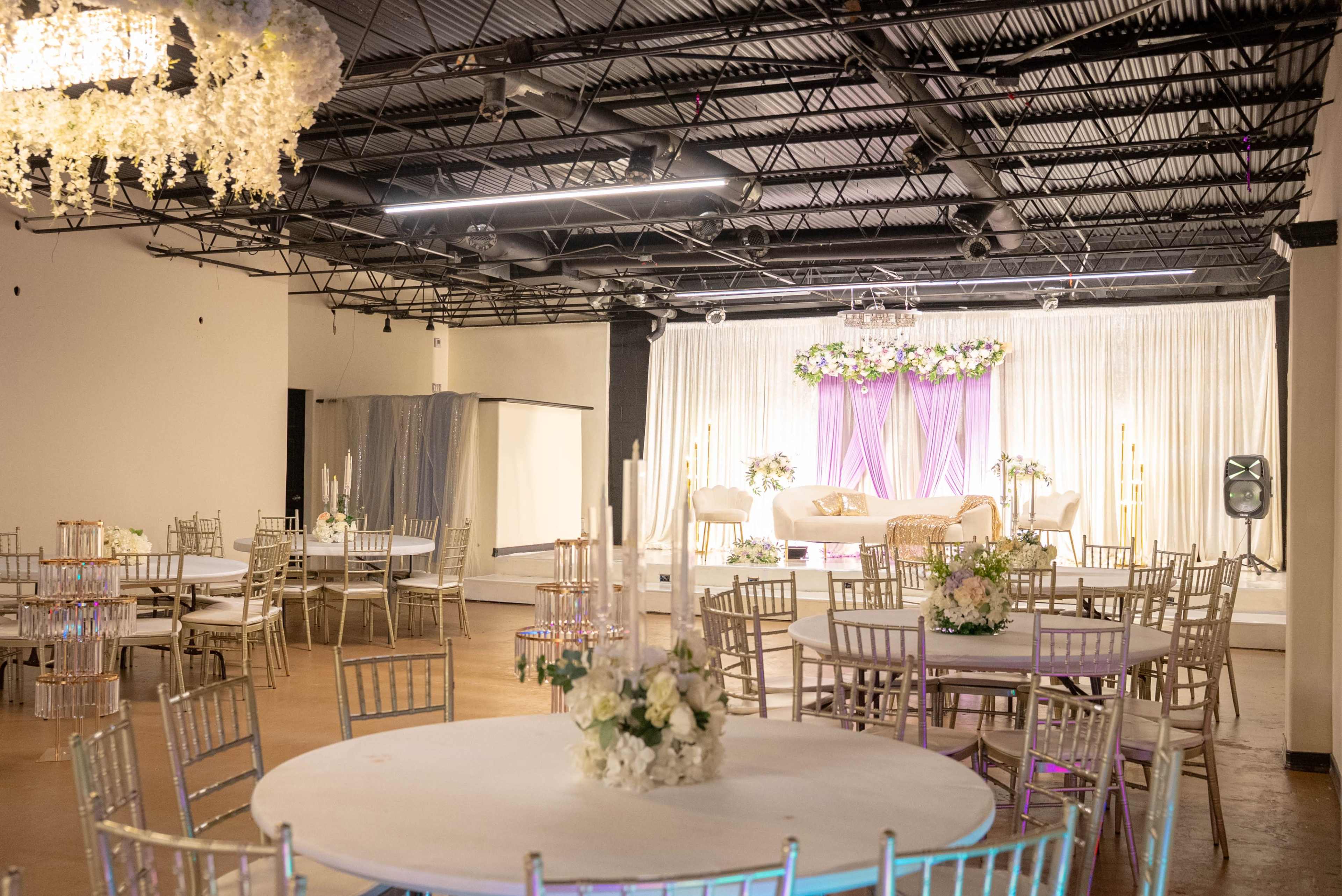 The image features a spacious event hall with round tables, gold chairs, and a decorated stage at the front draped in white and purple fabrics.