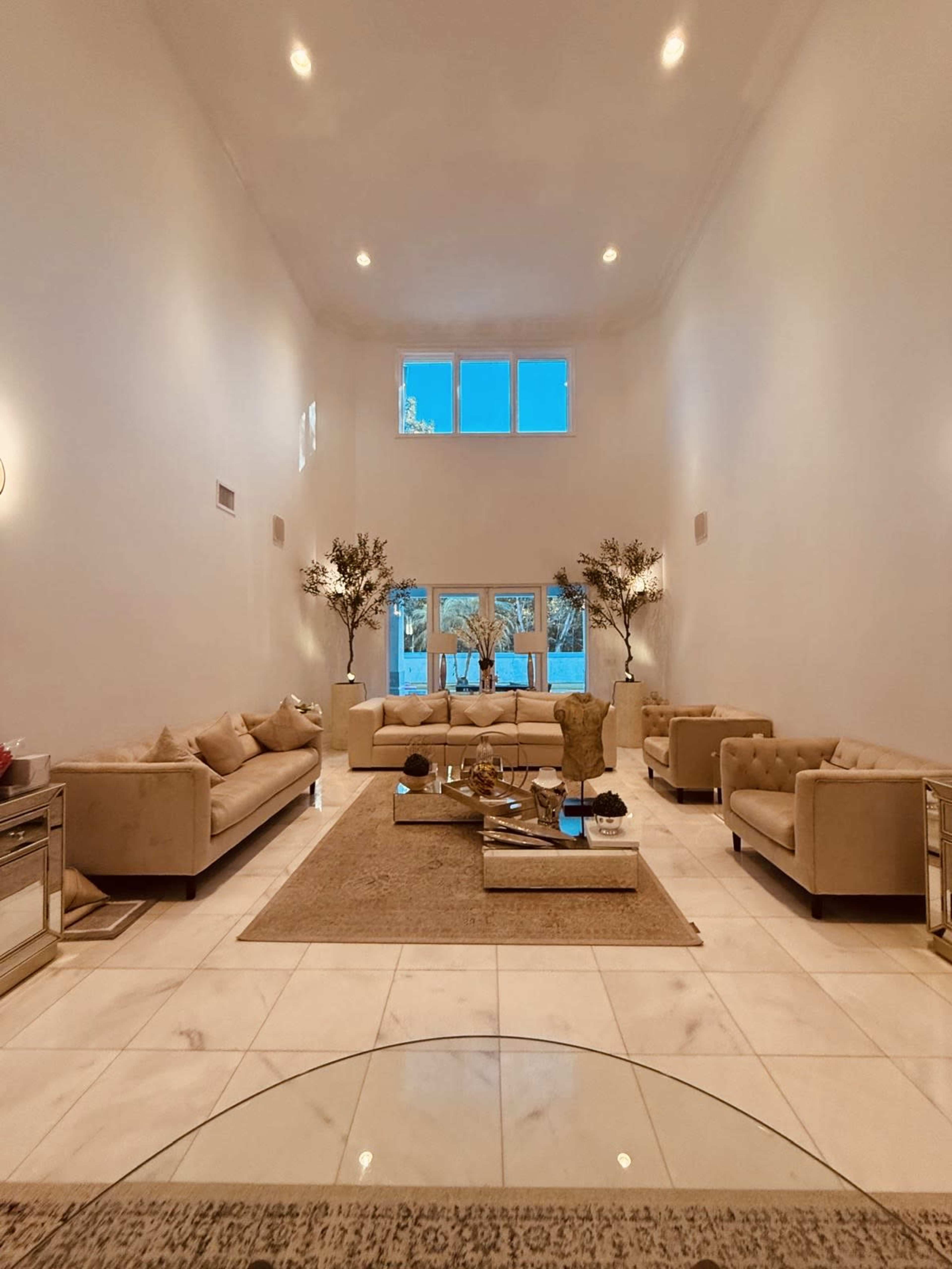 The image shows a spacious living room with beige sofas, a central coffee table, decorative plants, and large windows allowing natural light to enter.