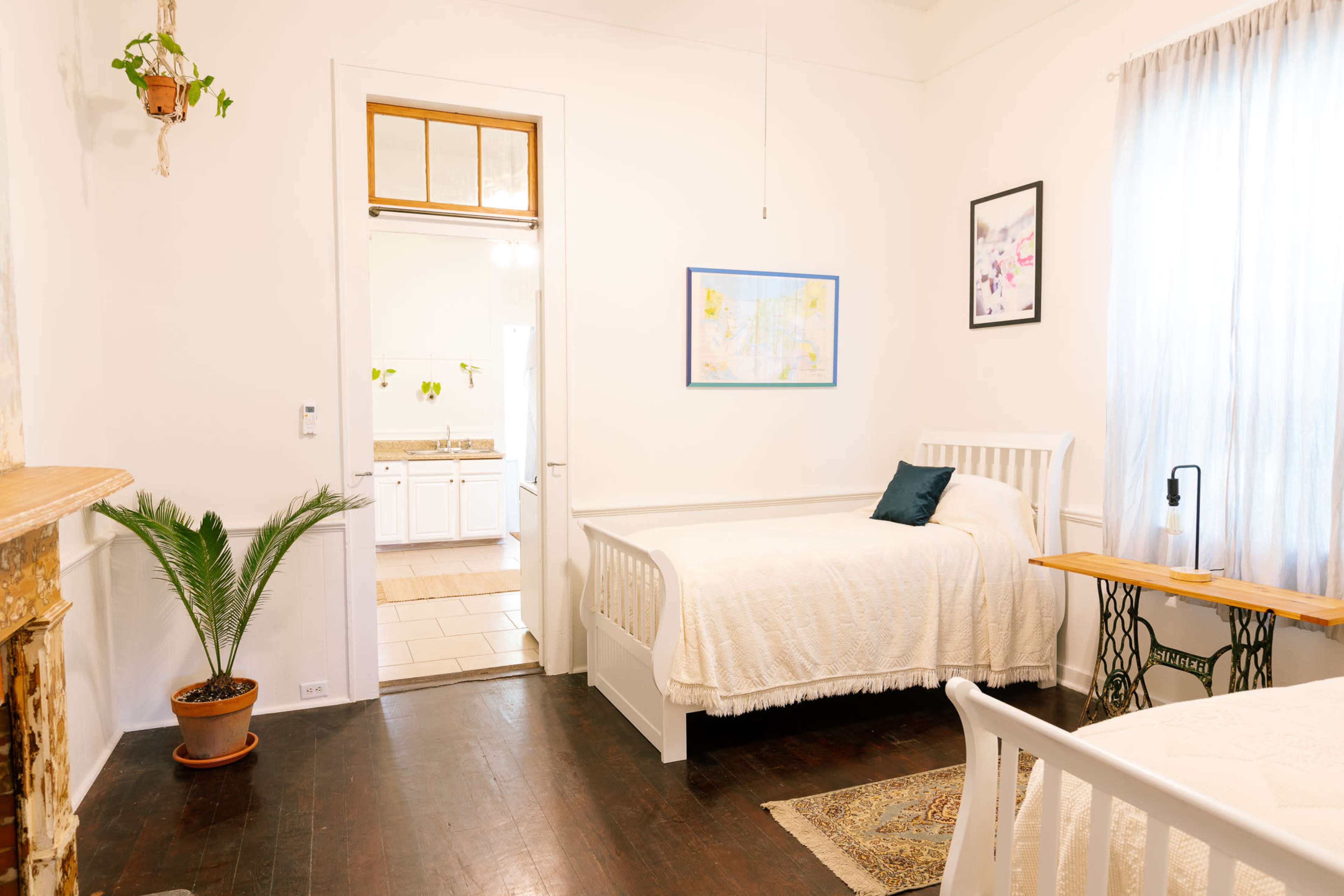 The image shows a bright and tidy bedroom featuring two beds, a small wooden table, and a door leading to a kitchenette area.