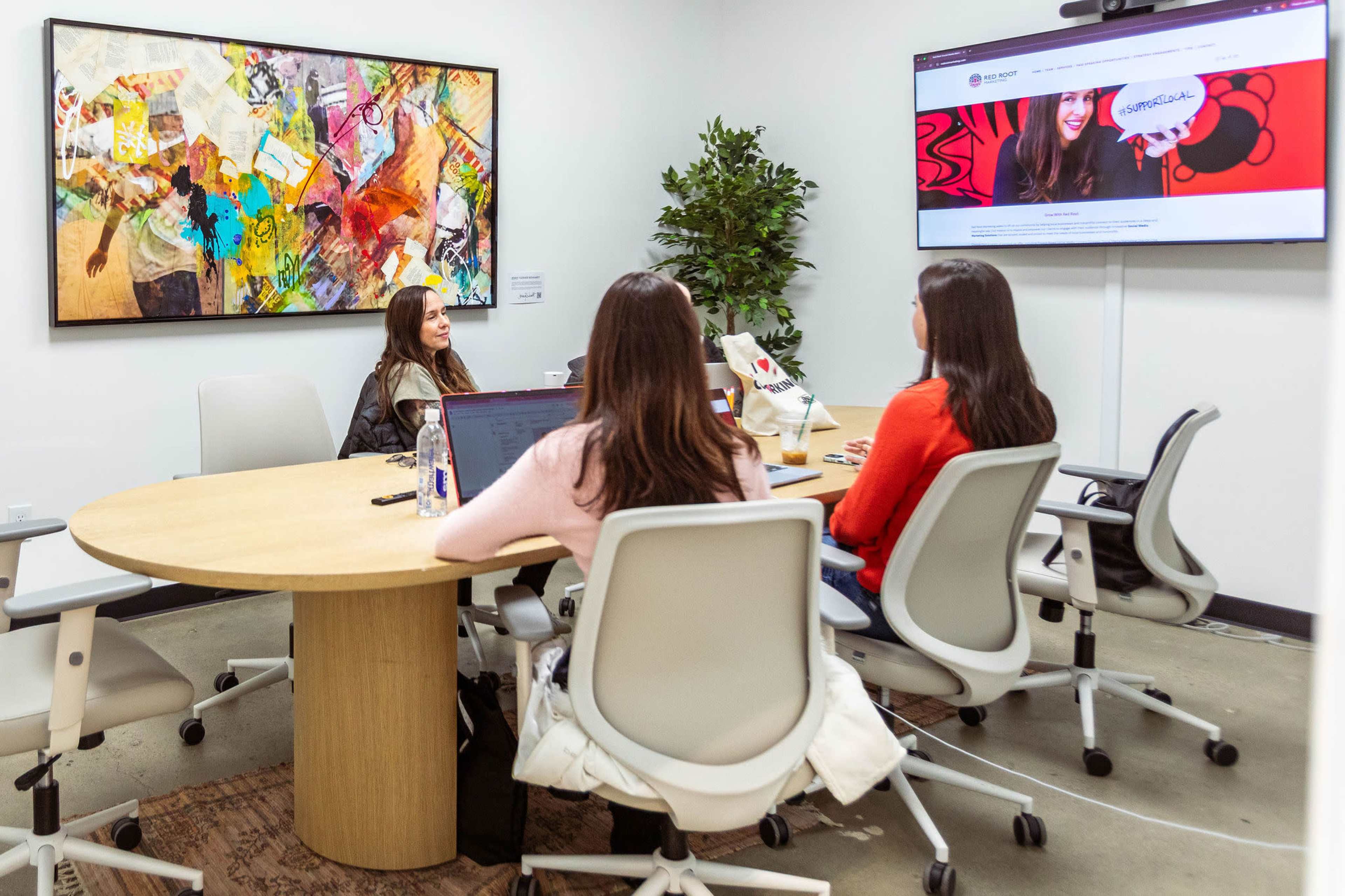 A group of four people is seated around a circular table in a meeting room, with an artwork on the wall and a large screen displaying a website.