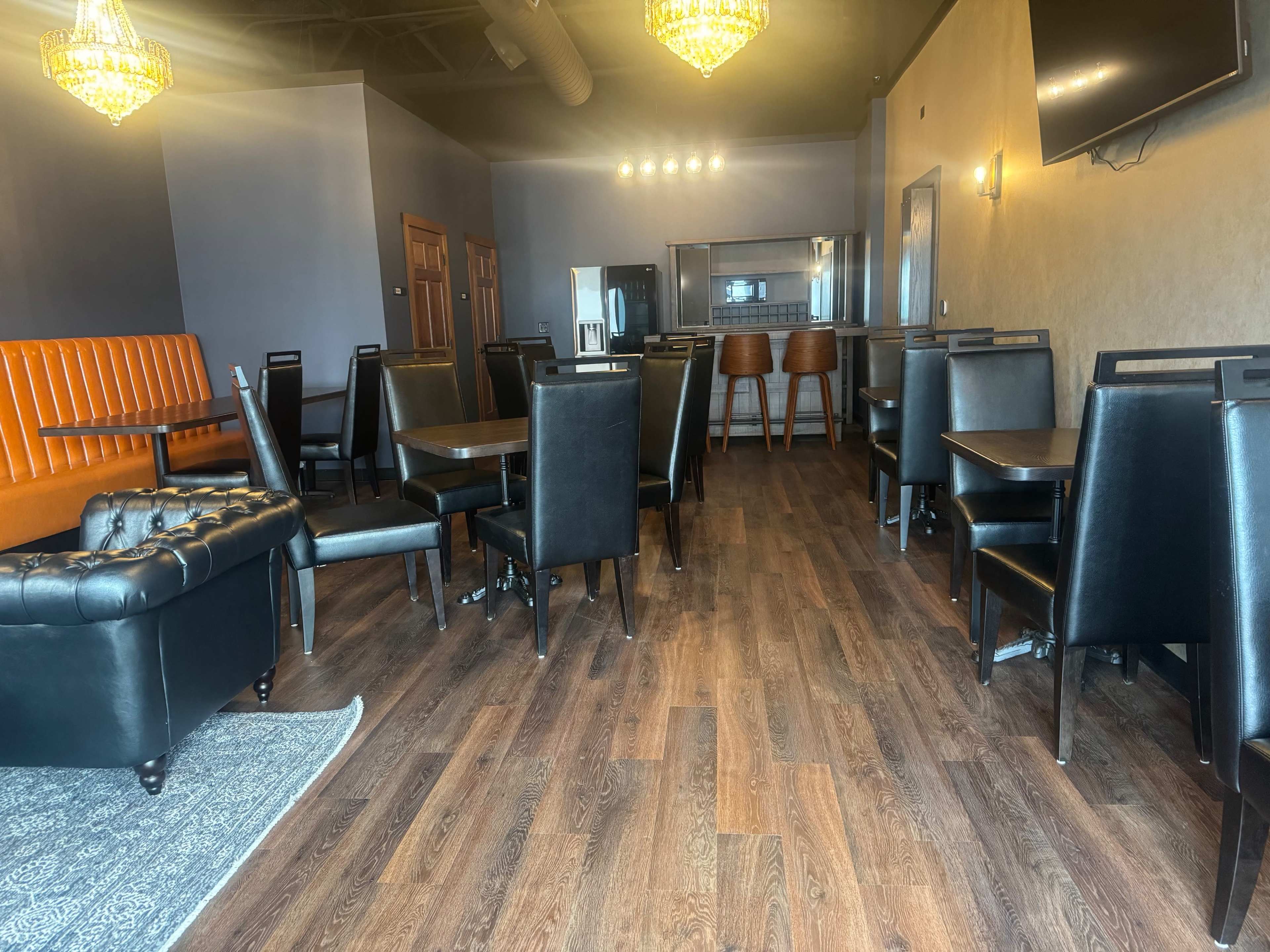 The image shows a modern dining area with black chairs at wooden tables, a brown leather sofa, and chandeliers overhead.