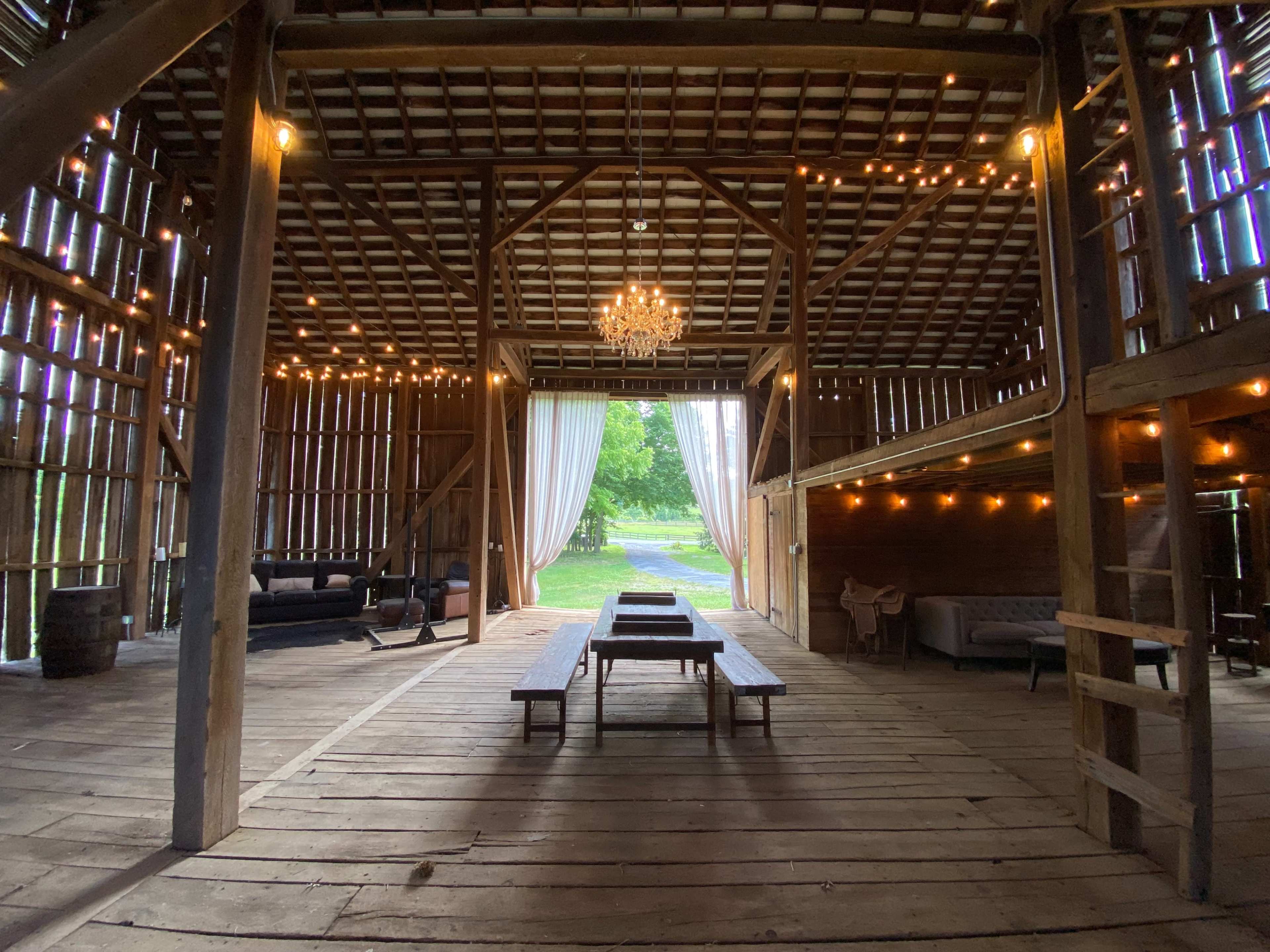 The interior of a rustic barn featuring wooden floors, string lights, a chandelier, and furniture arranged around a central table.