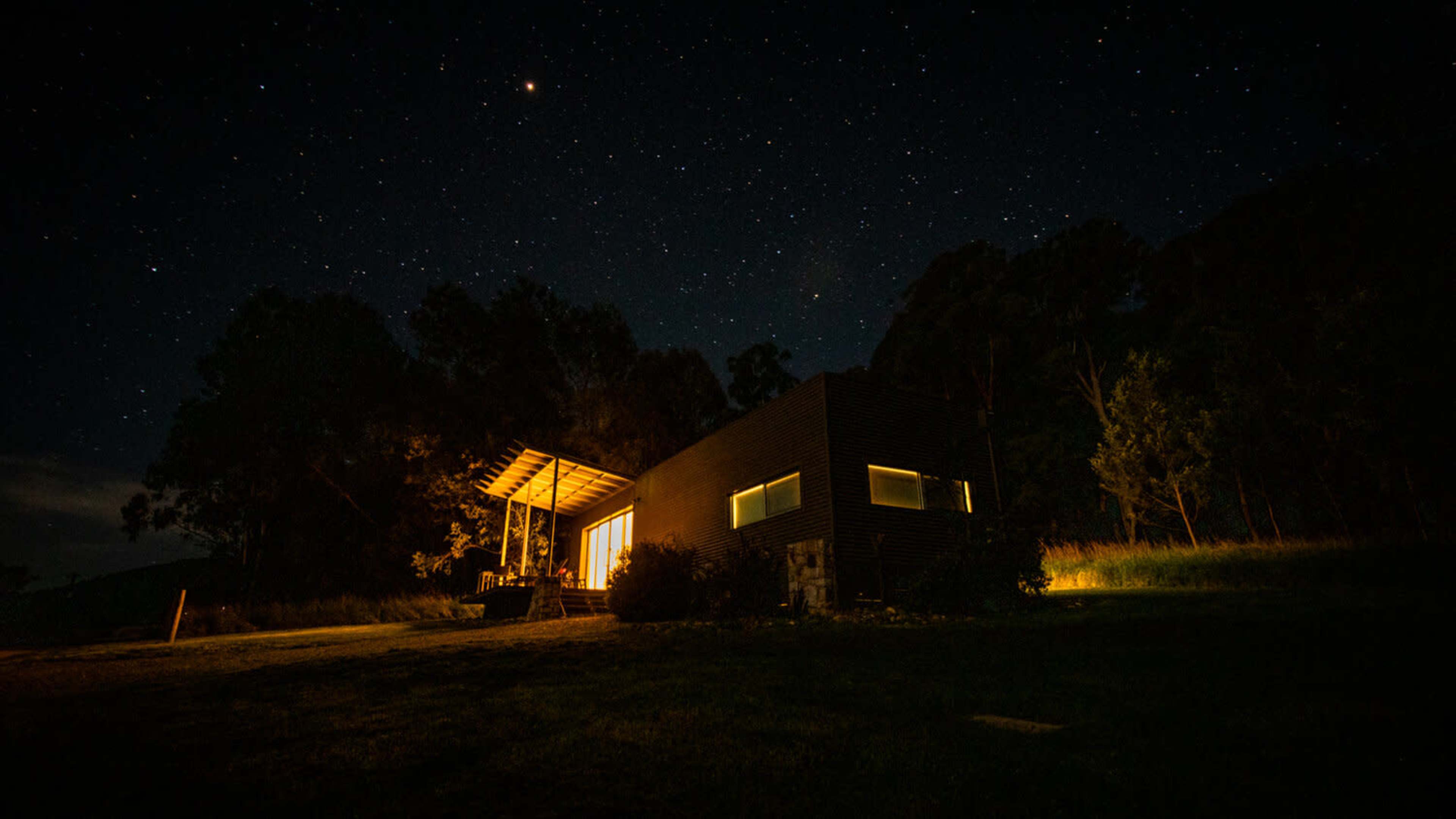 A modern house with illuminated windows stands in a dark landscape under a starry night sky.