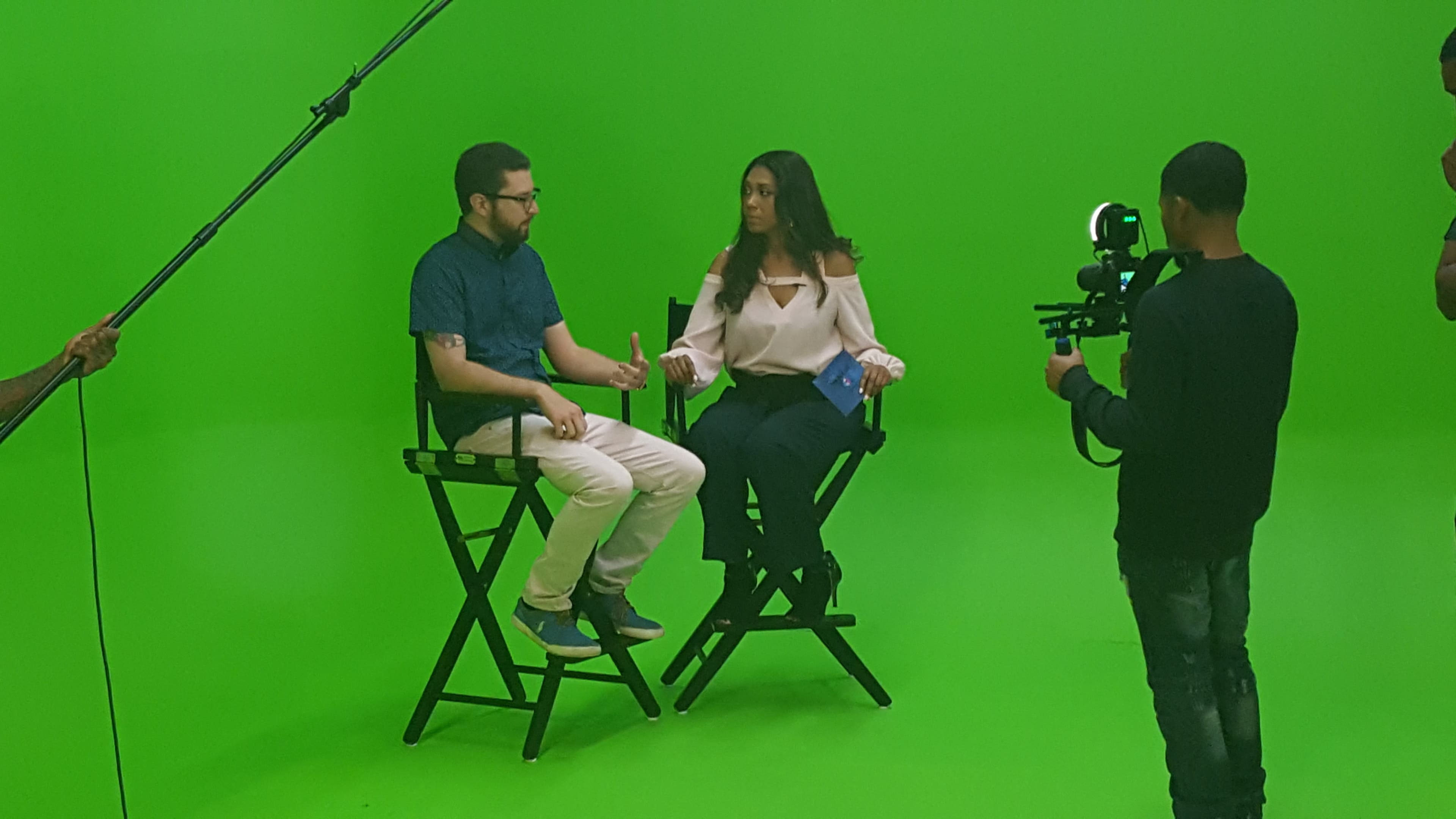 A man and a woman sit in director's chairs on a green screen set while a camera operator records the scene.
