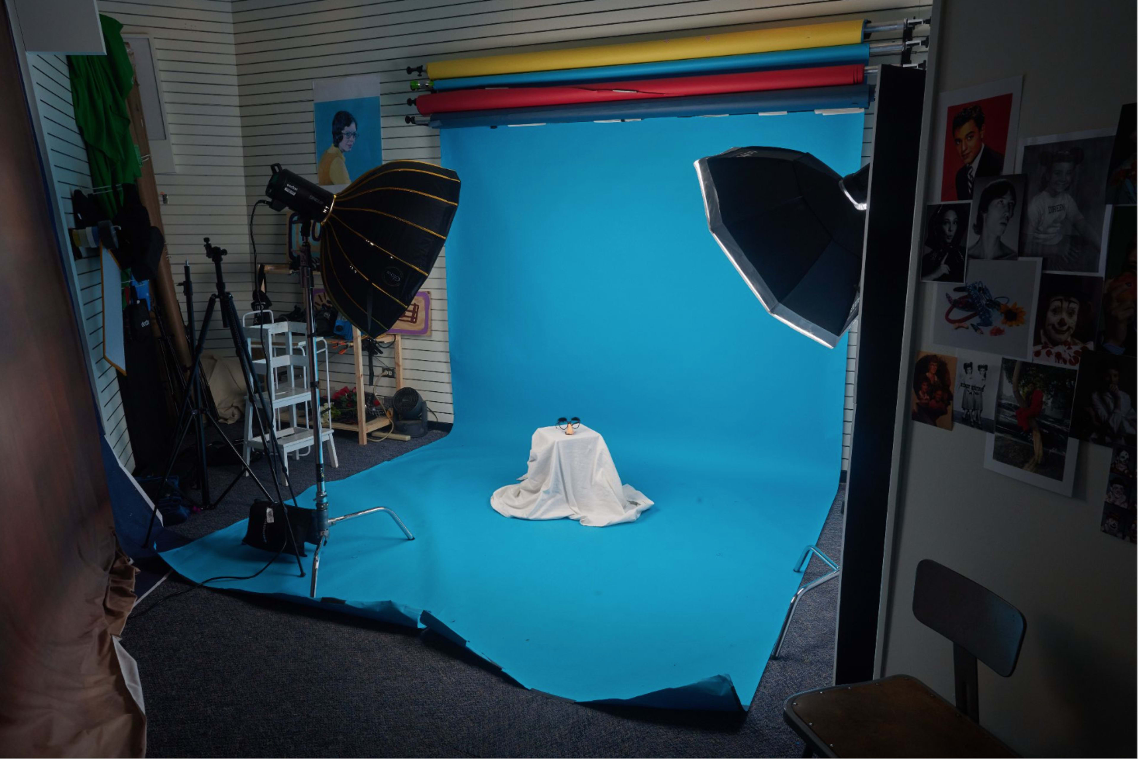 The image shows a photography studio with a blue backdrop, two softbox lights, a table covered with a white cloth, and various photography equipment arranged around the space.