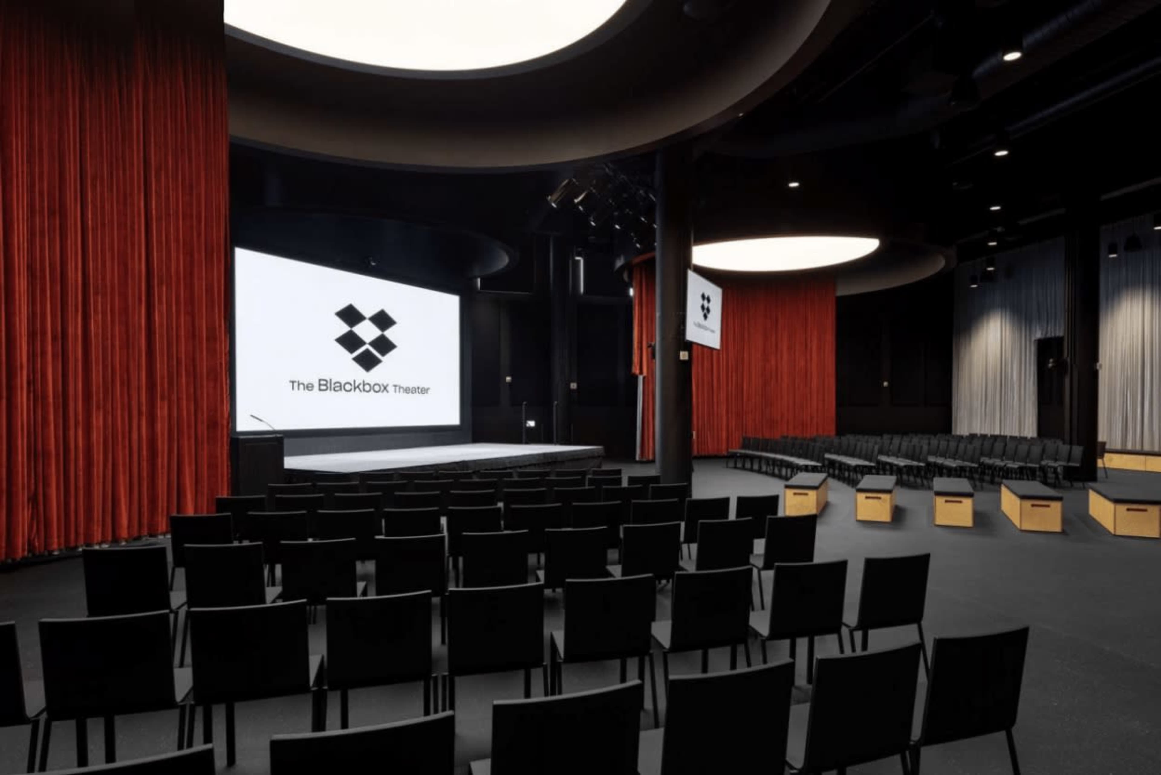 A modern theater space with rows of black chairs facing a large screen, flanked by red curtains.