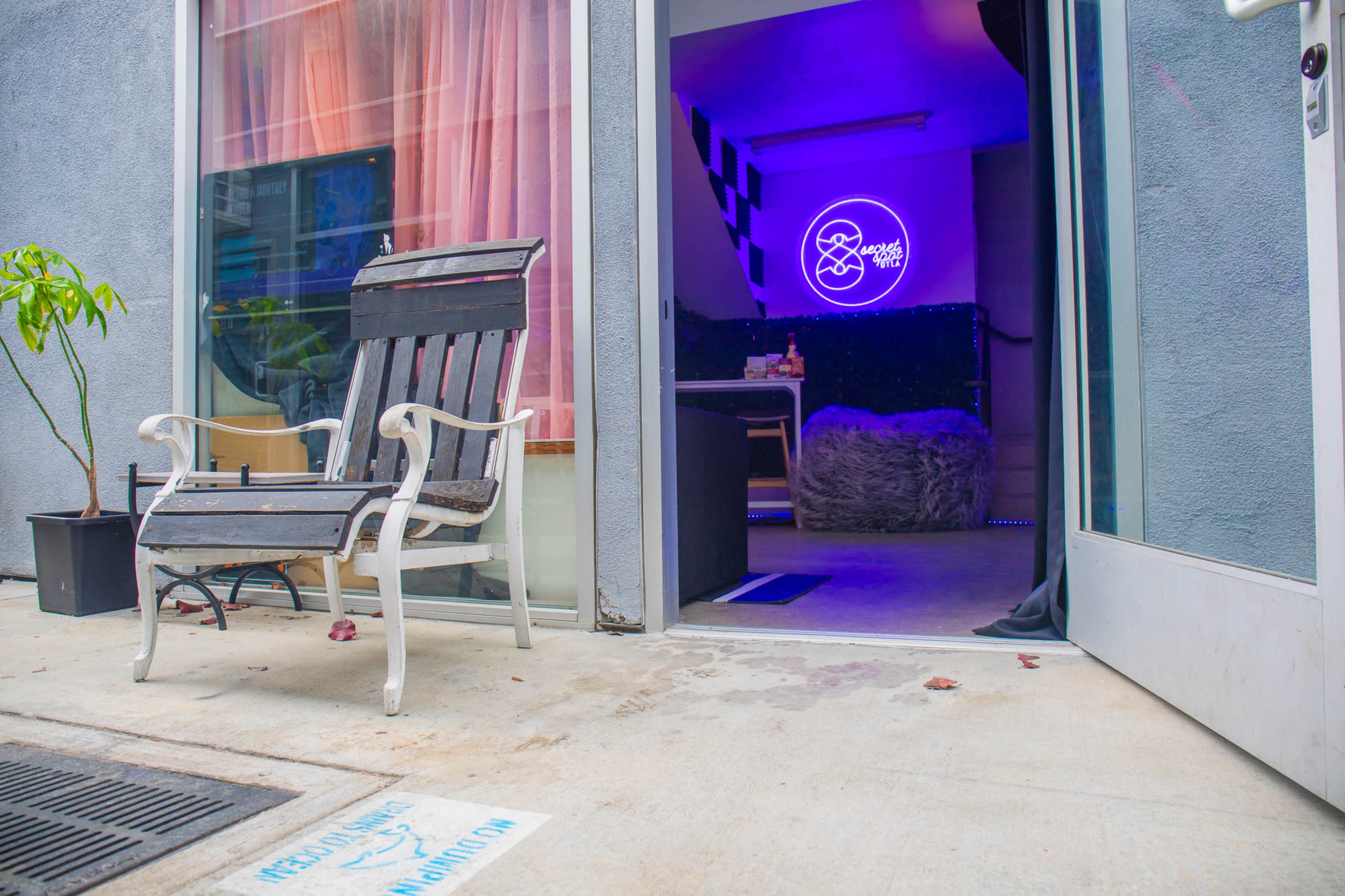 A black and white striped chair sits on a concrete patio in front of an open doorway leading to an interior space illuminated by a purple neon sign.