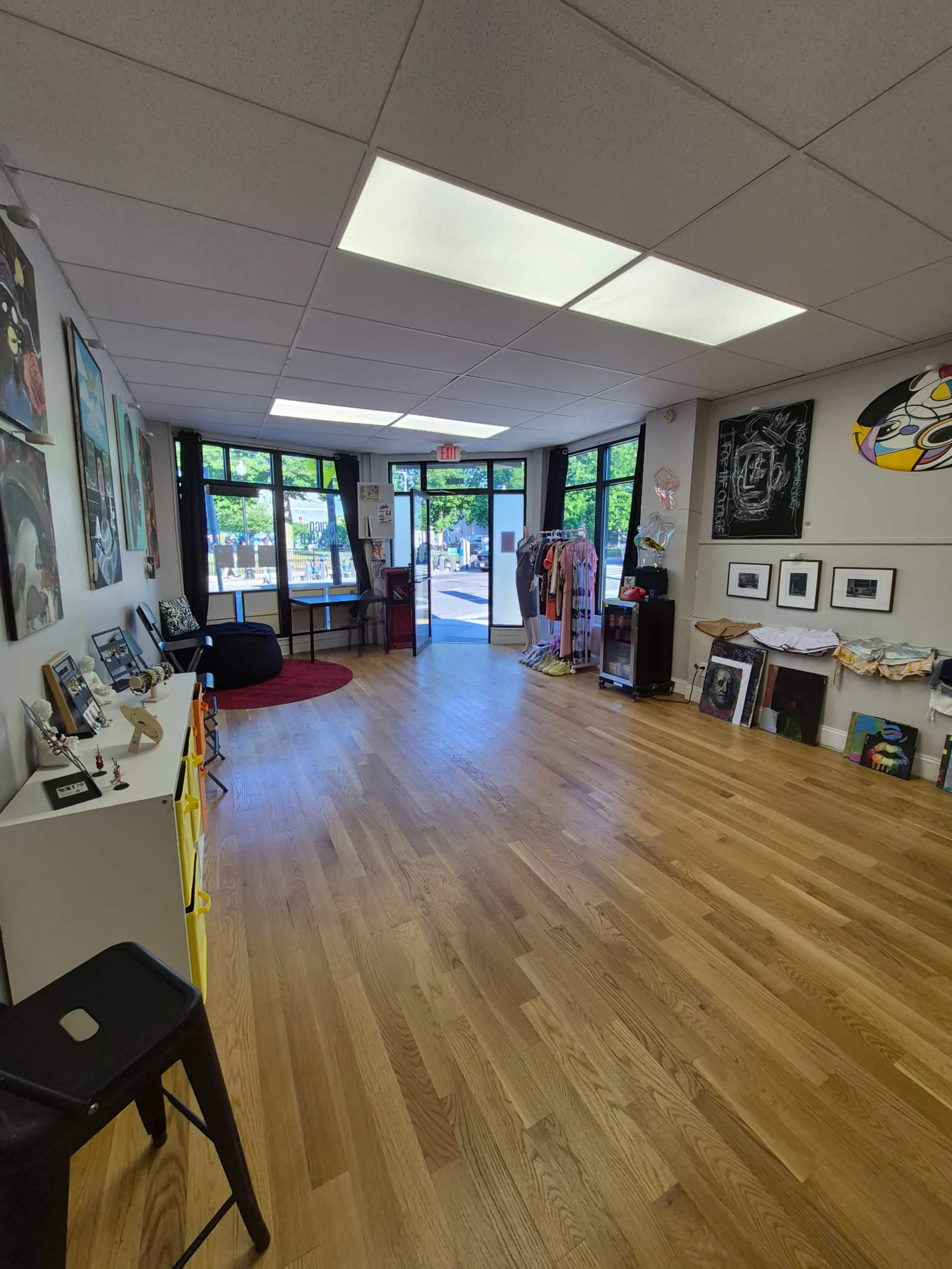 The image shows an empty art gallery with wooden flooring, large windows, and an open entrance leading to an outdoor area.