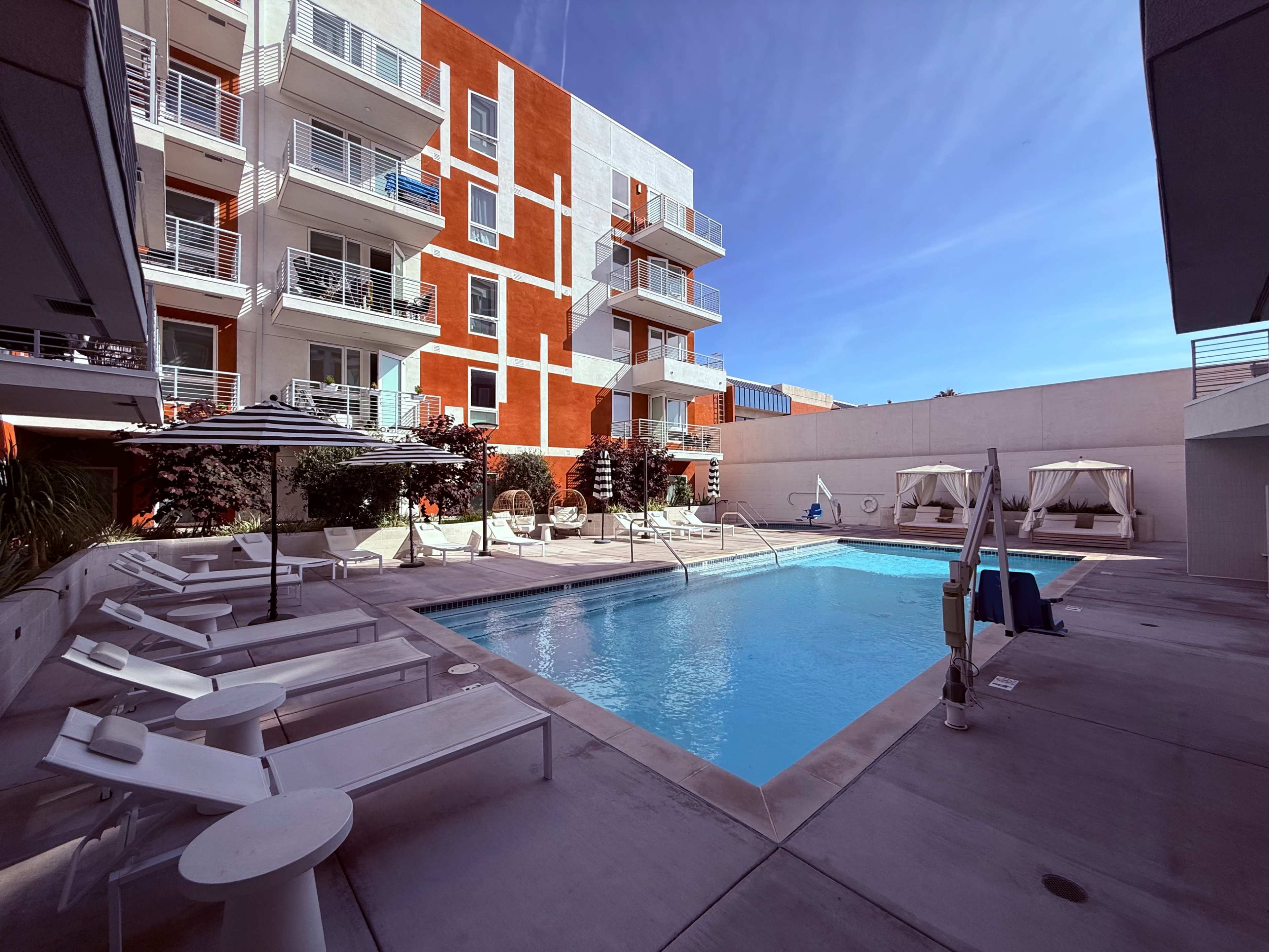 The image shows a rectangular swimming pool surrounded by lounge chairs and cabanas, located in an outdoor area of a modern apartment complex with bright red and white walls.