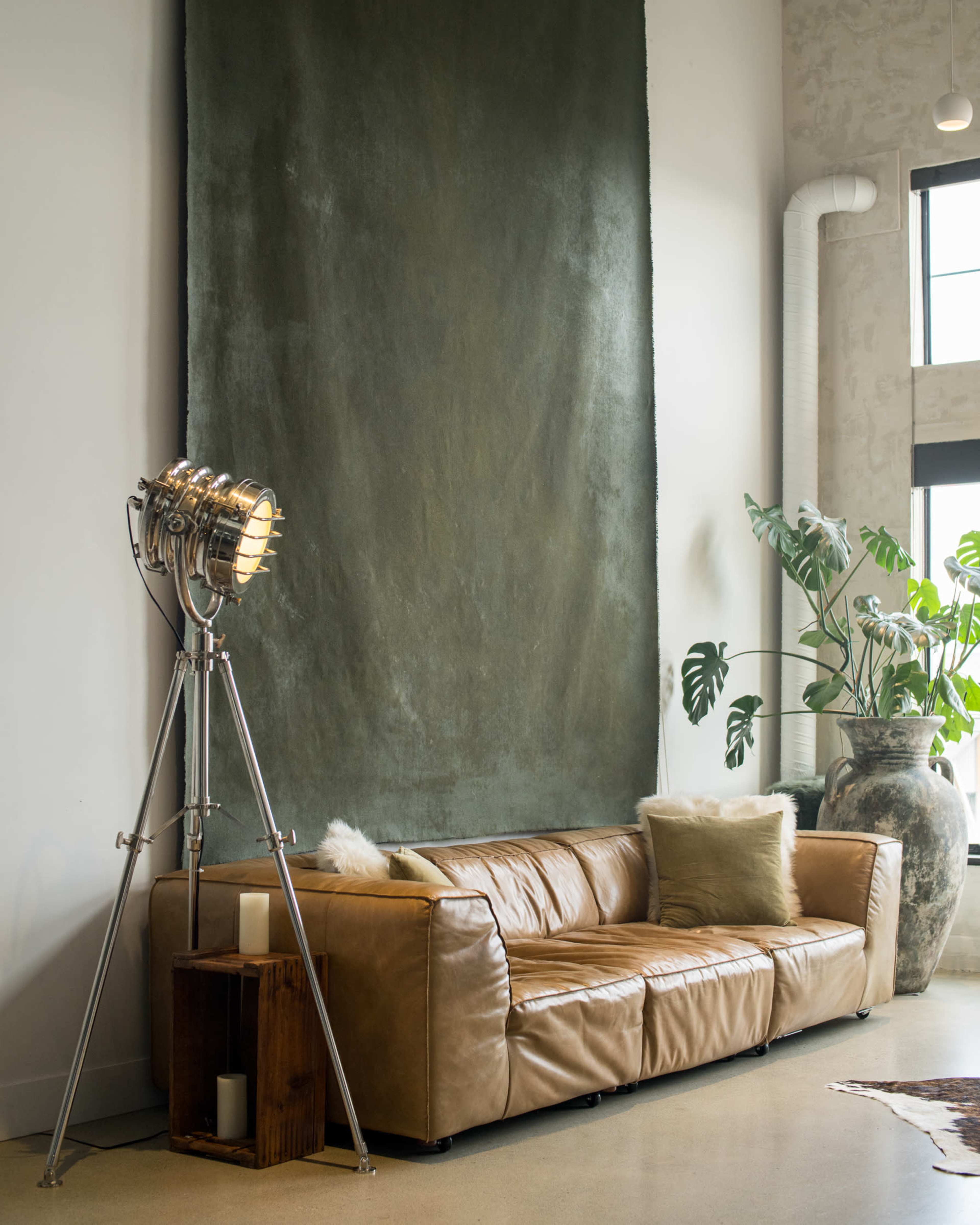 A brown leather sofa sits against a textured green wall, next to a tall vase with leafy plants and a vintage-style floor lamp.
