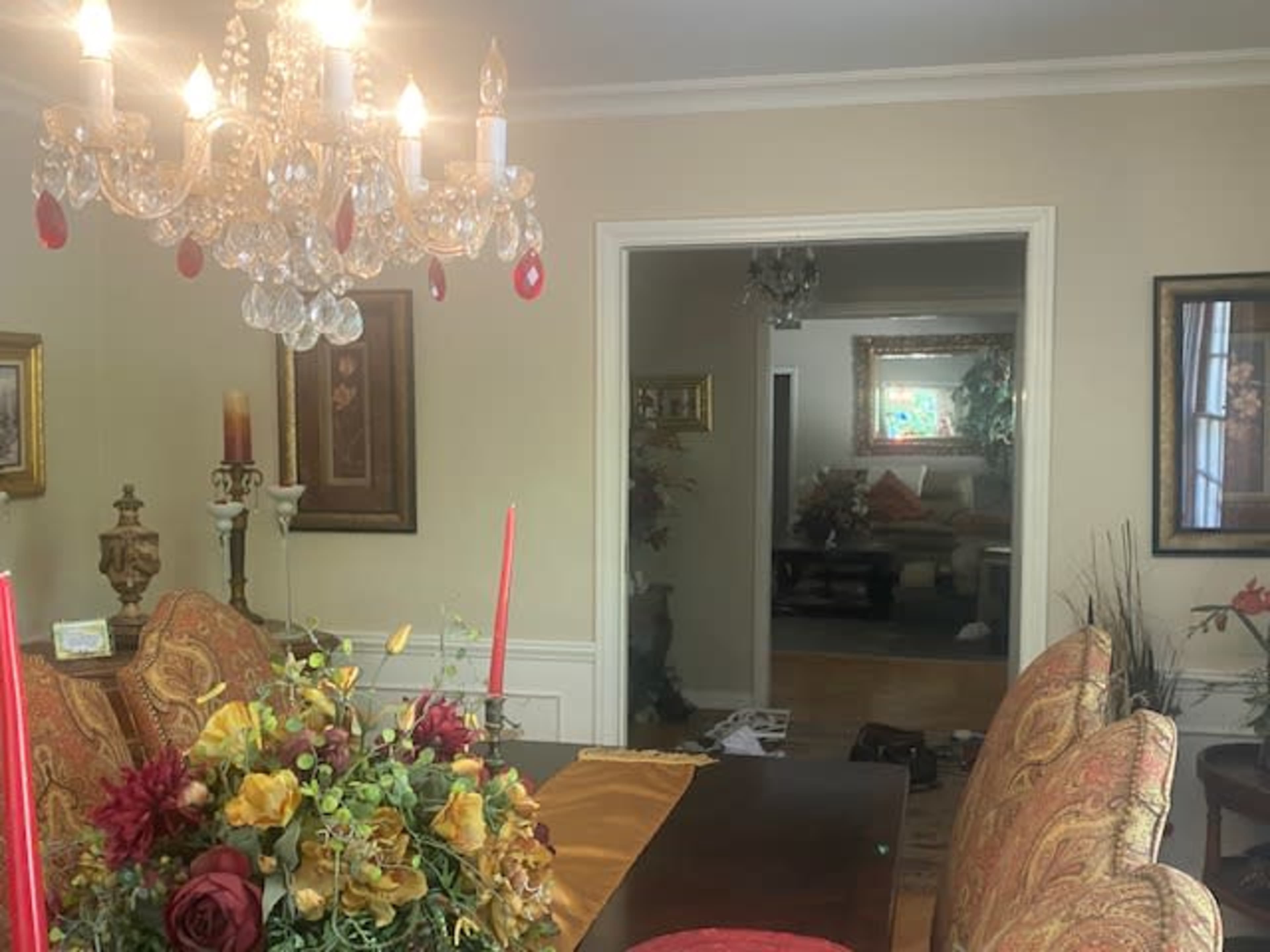 A formal dining room features a large table adorned with decorative flowers and candles, illuminated by a chandelier, with a doorway leading to a living area in the background.