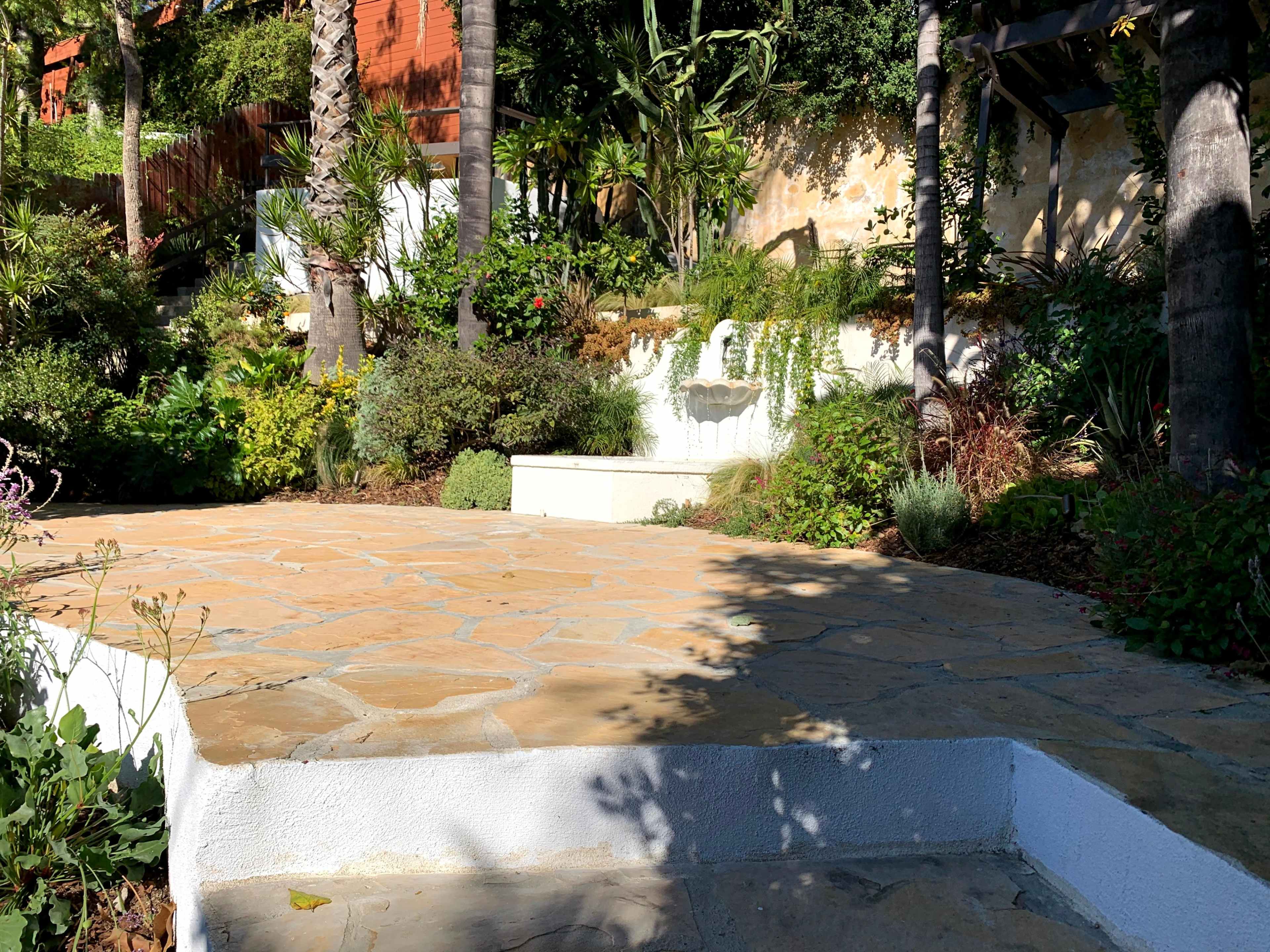 The image shows a landscaped garden with a stone pathway, featuring various plants and a small waterfall against a textured wall.