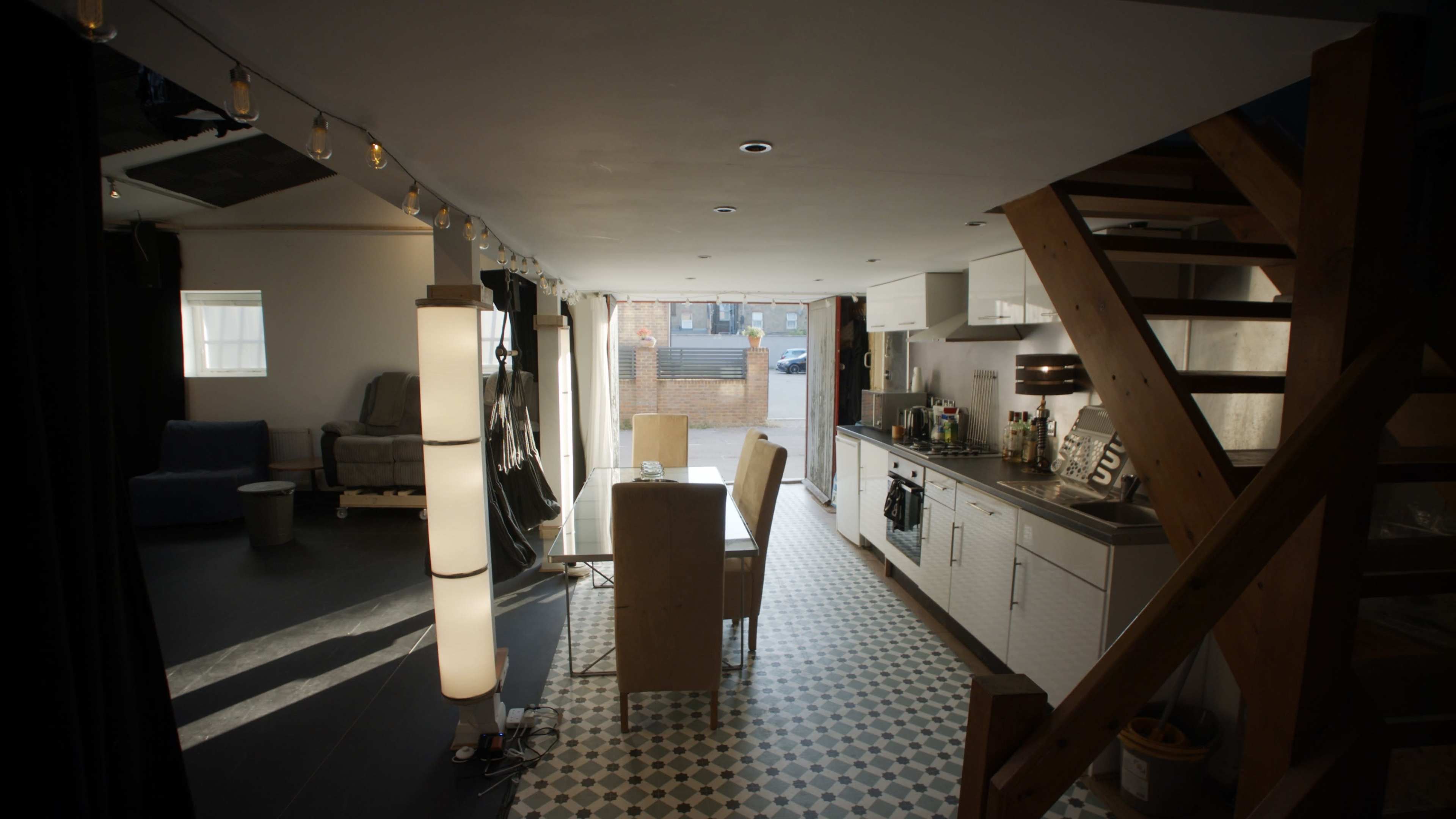 A modern kitchen and dining area with a table and chairs, connected to a staircase and featuring large windows that open to an outdoor space.
