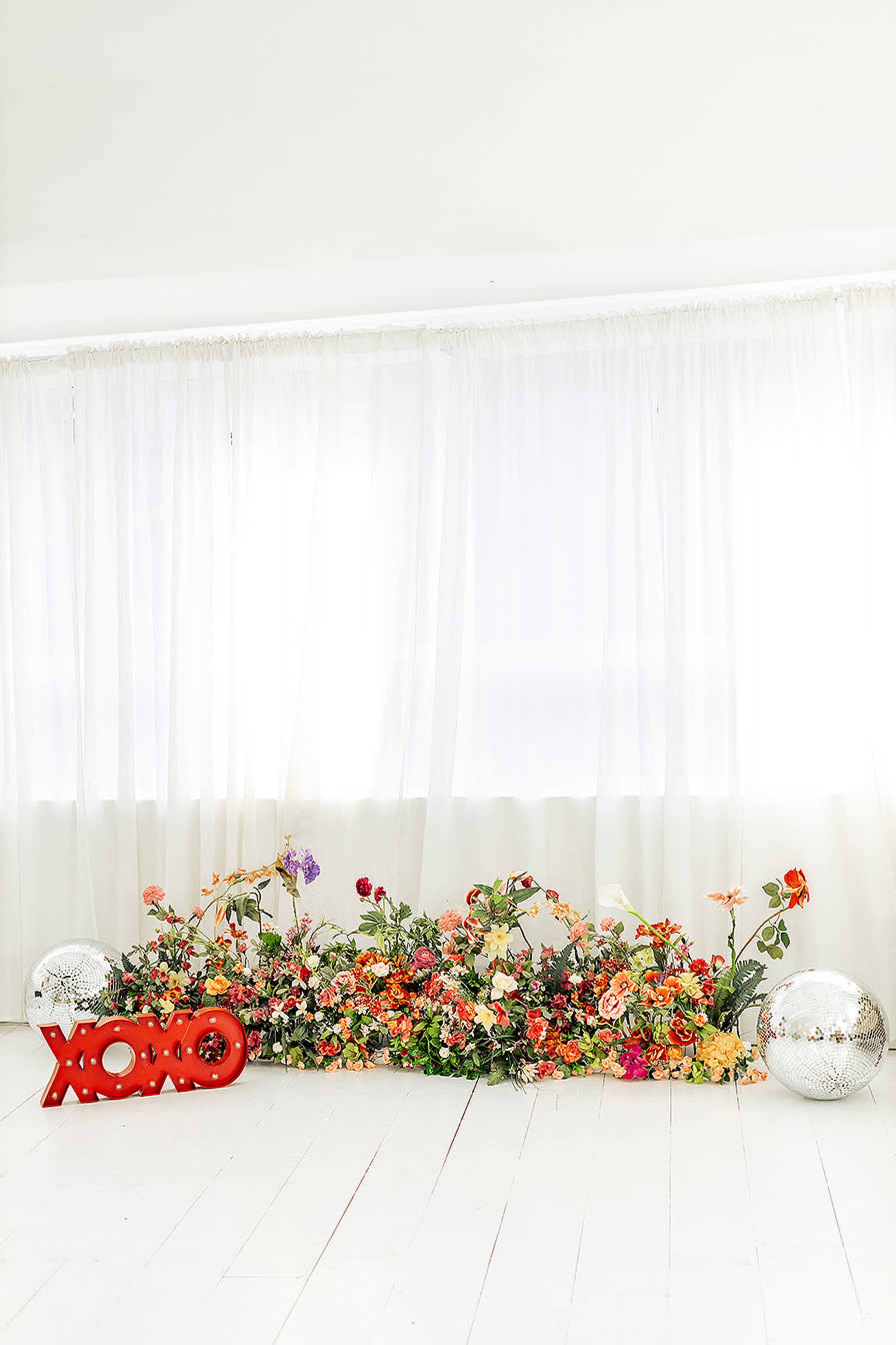 A bright interior scene features a row of colorful flowers along the floor with a red "XOXO" sign and two reflective disco balls in front of sheer white curtains.