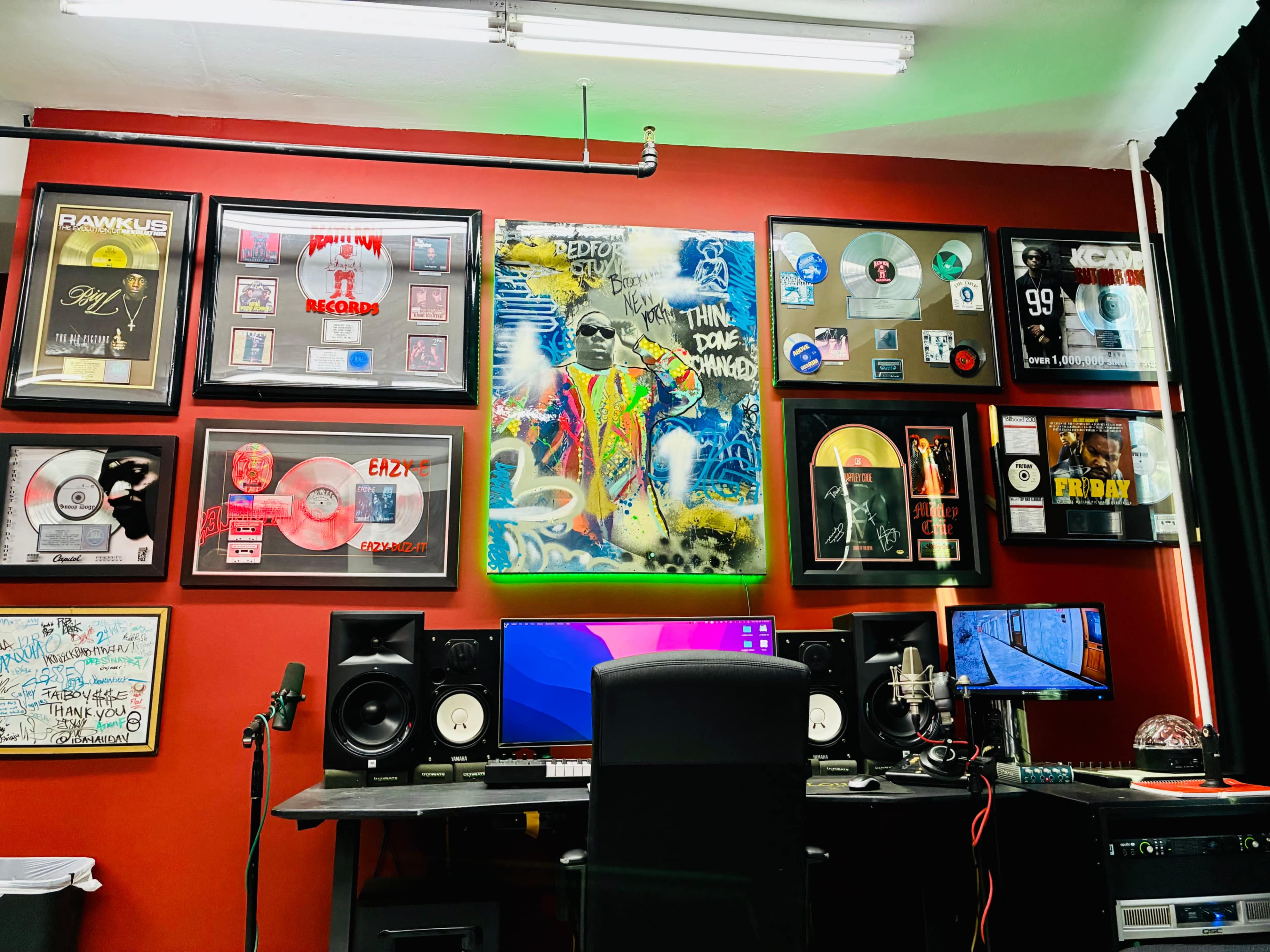 The image shows a well-decorated music studio featuring a desk with audio equipment, two speakers, and multiple gold and platinum record plaques displayed on red walls.