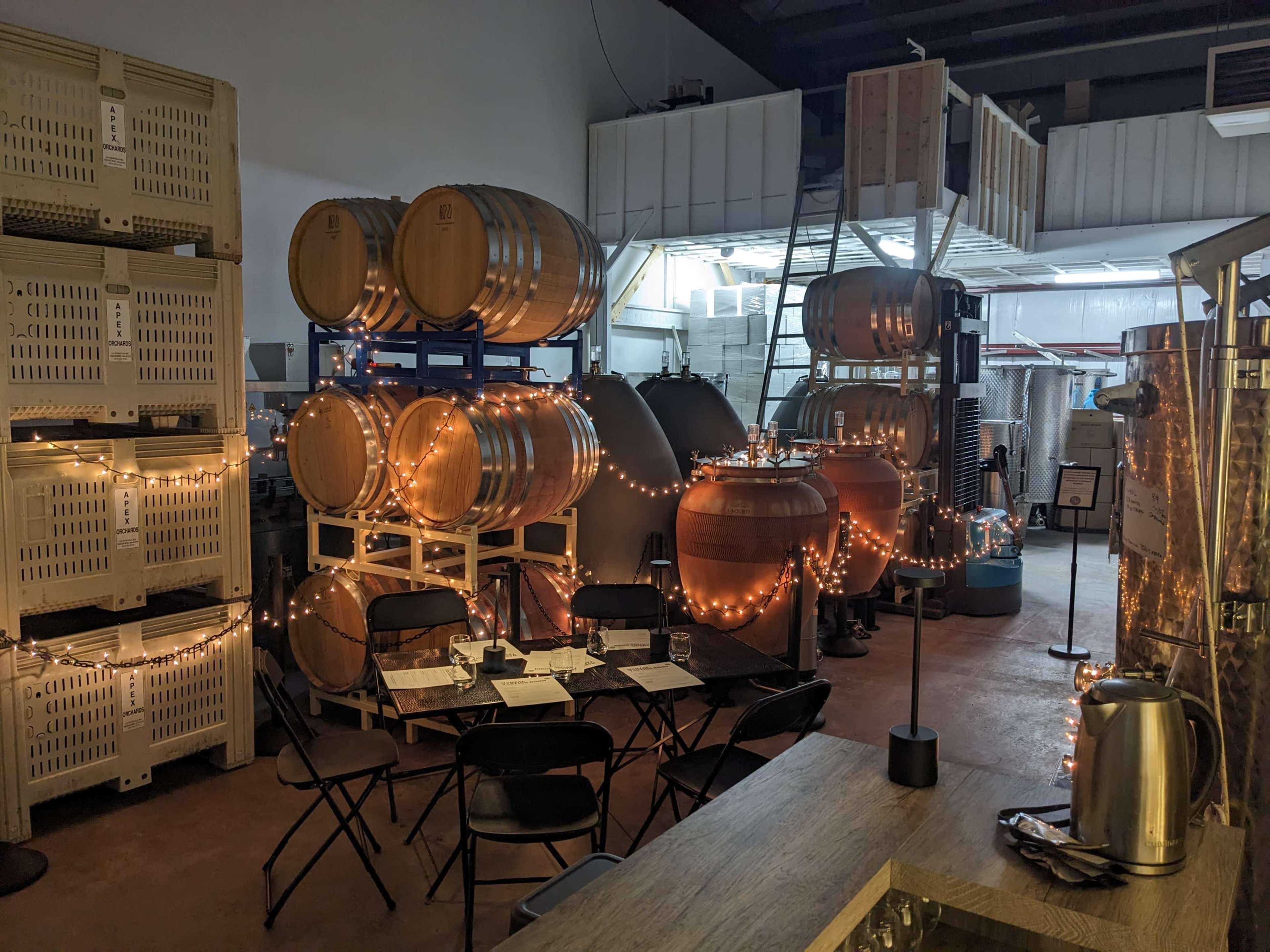 A wine production facility featuring wooden barrels, stainless steel tanks, and a small seating area decorated with string lights.