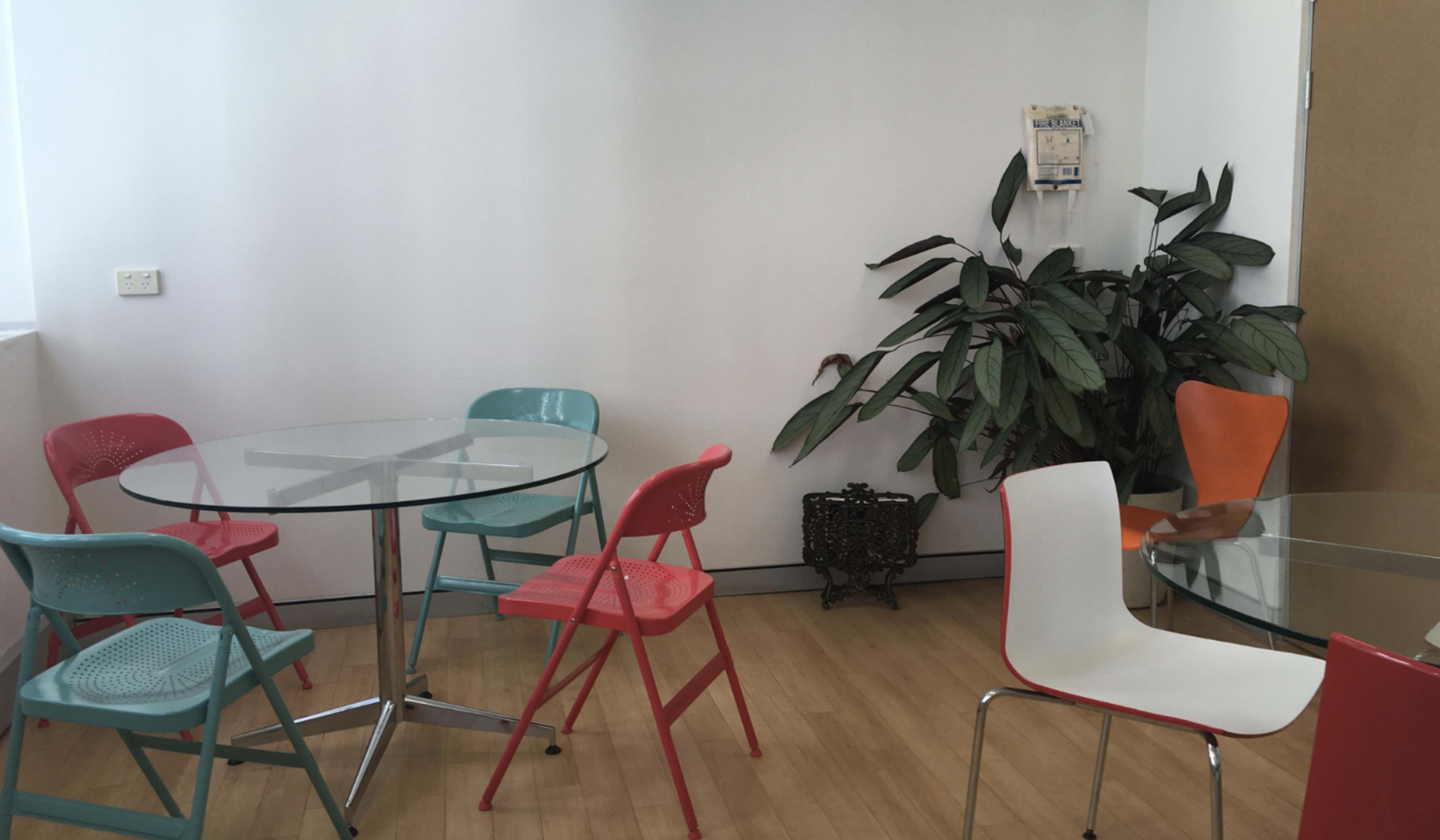 The image shows a modern room with glass tables and colorful chairs arranged around them, alongside a large potted plant.