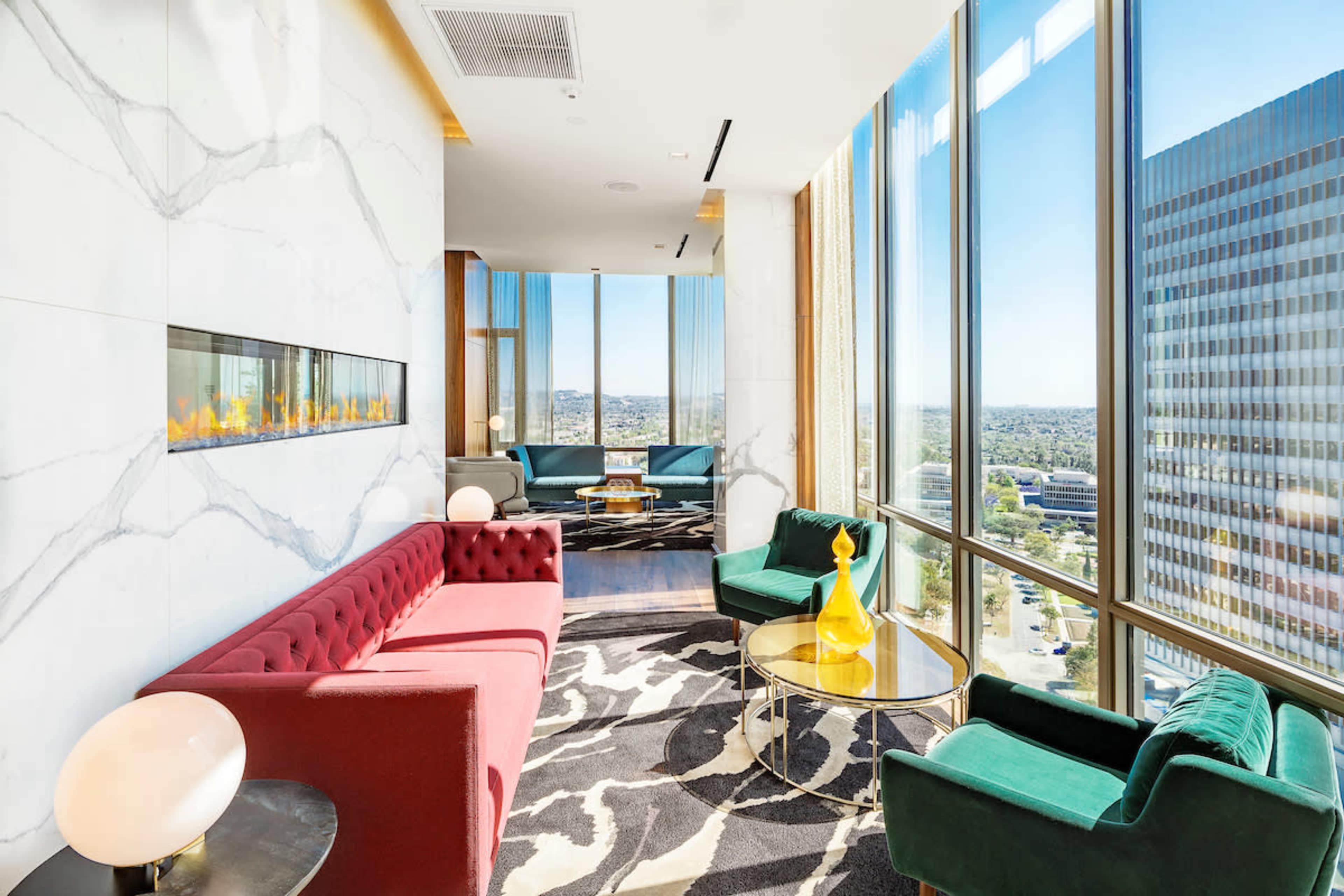 The image shows a modern lounge area with large windows, featuring a red tufted sofa, green armchairs, and a glass coffee table, overlooking a cityscape.