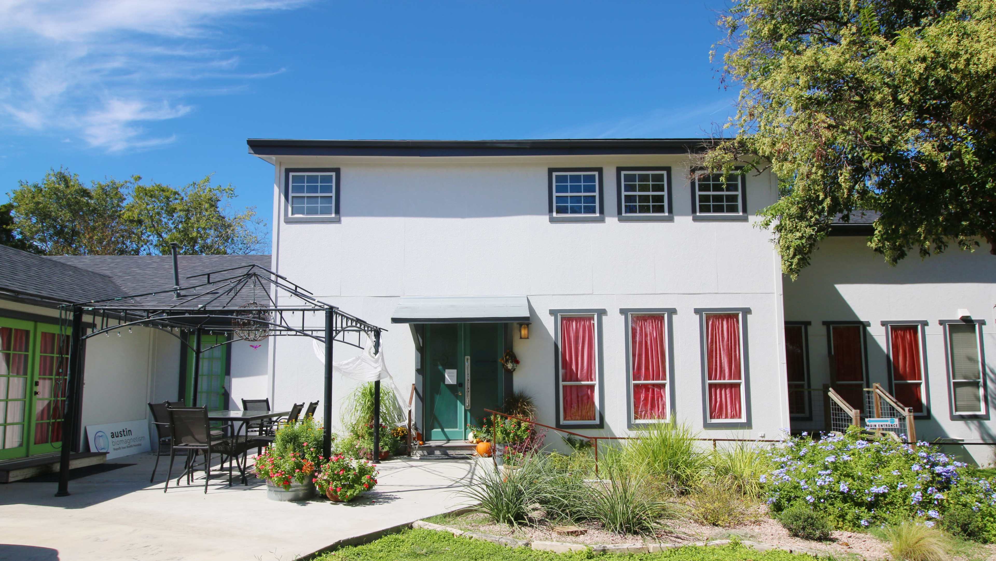The image shows a two-story building with a green door and large windows, surrounded by greenery and a seating area under a gazebo.