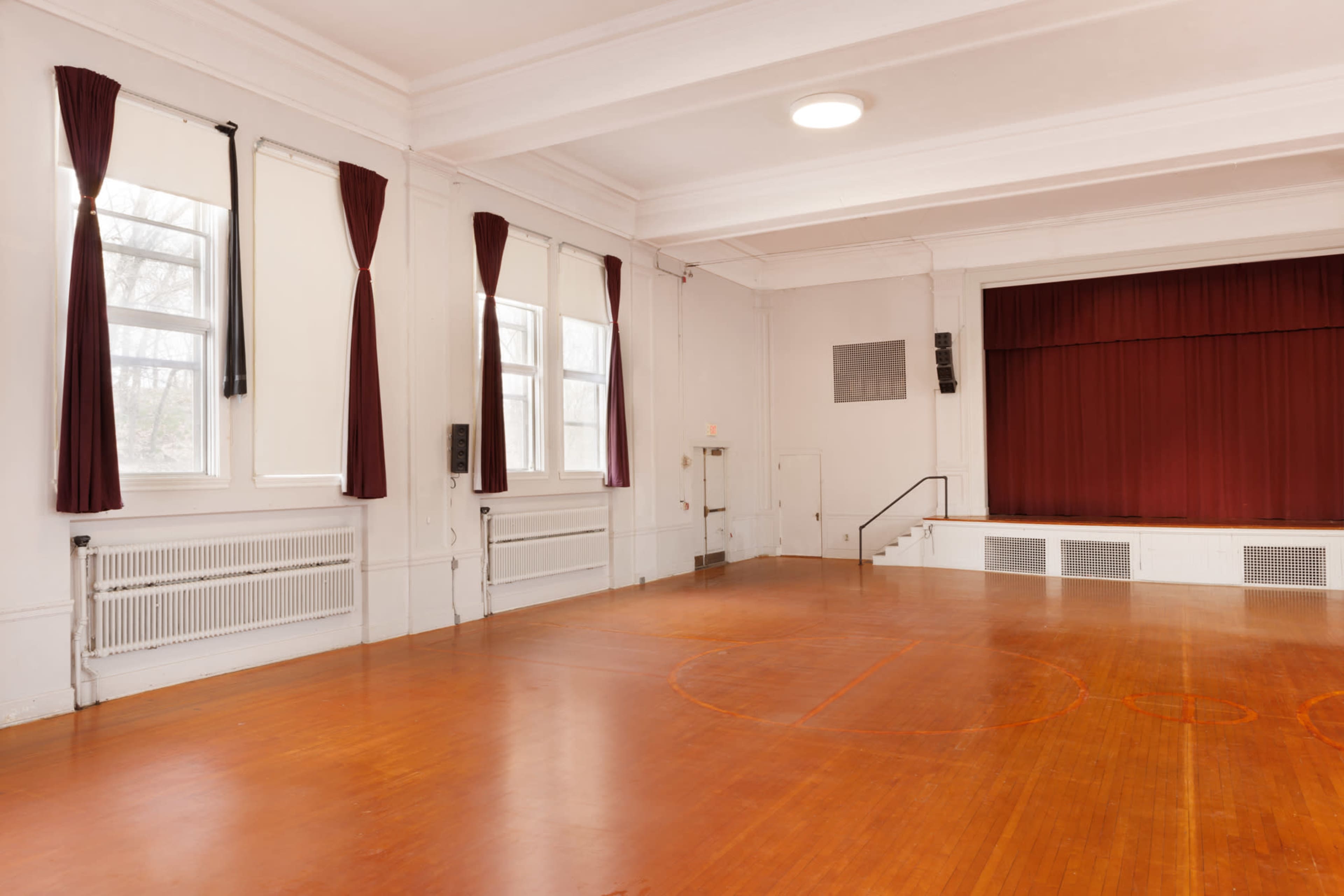 An empty gymnasium with wooden flooring, large windows, and a stage with red curtains at one end.