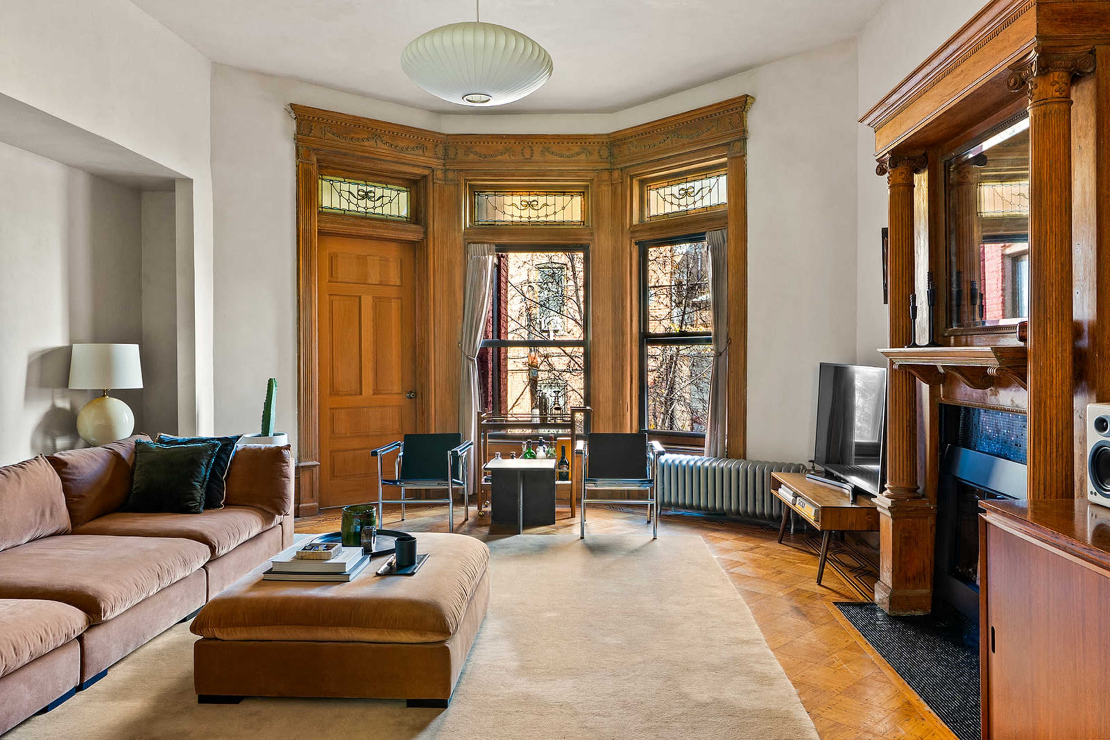 The room features a large, L-shaped sofa, a coffee table, and a wooden entertainment unit, with natural light streaming through the tall windows framed by intricate woodwork.