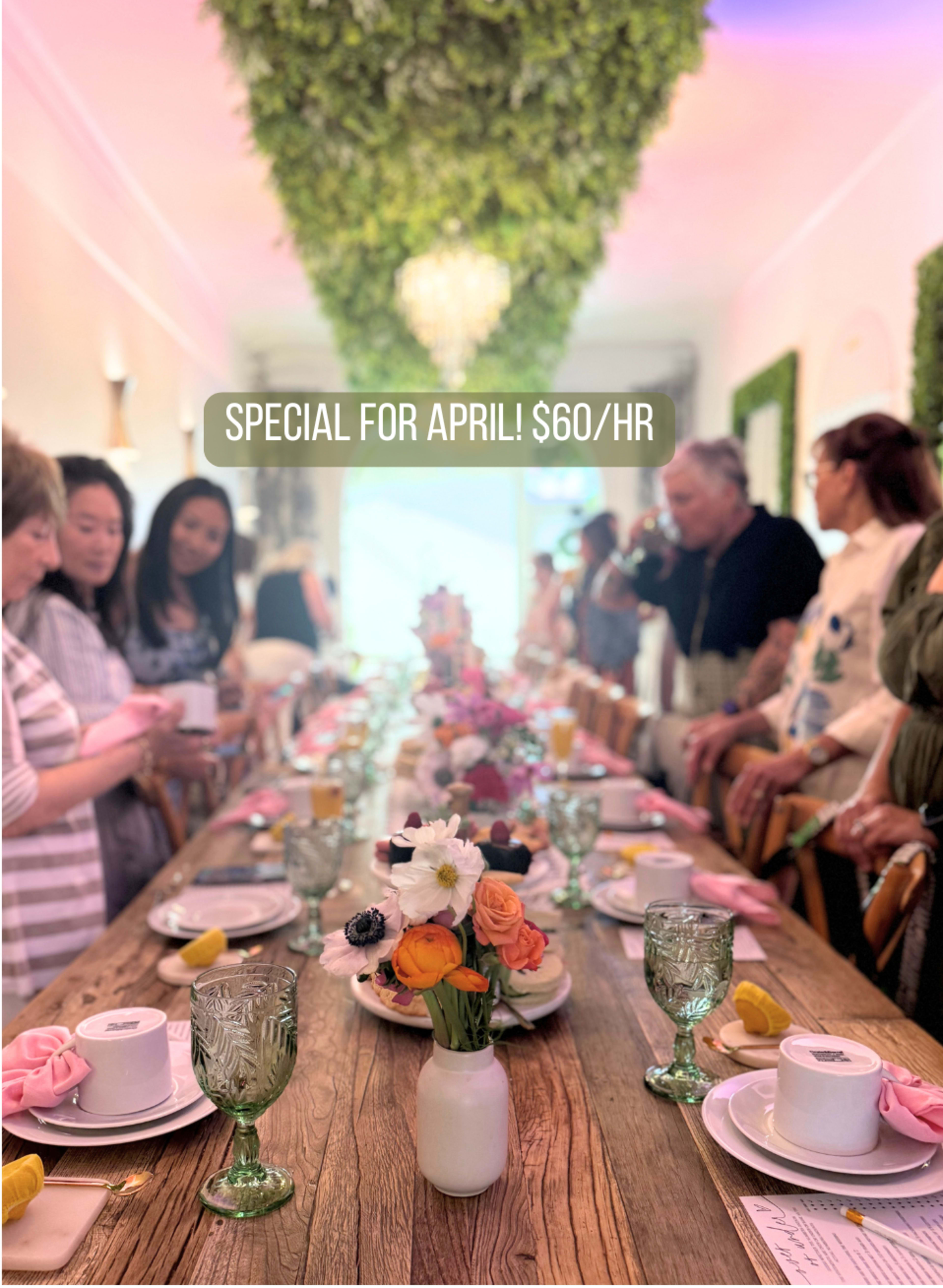 A long dining table is set with floral arrangements and decorative place settings, while people gather around in a bright room adorned with greenery.