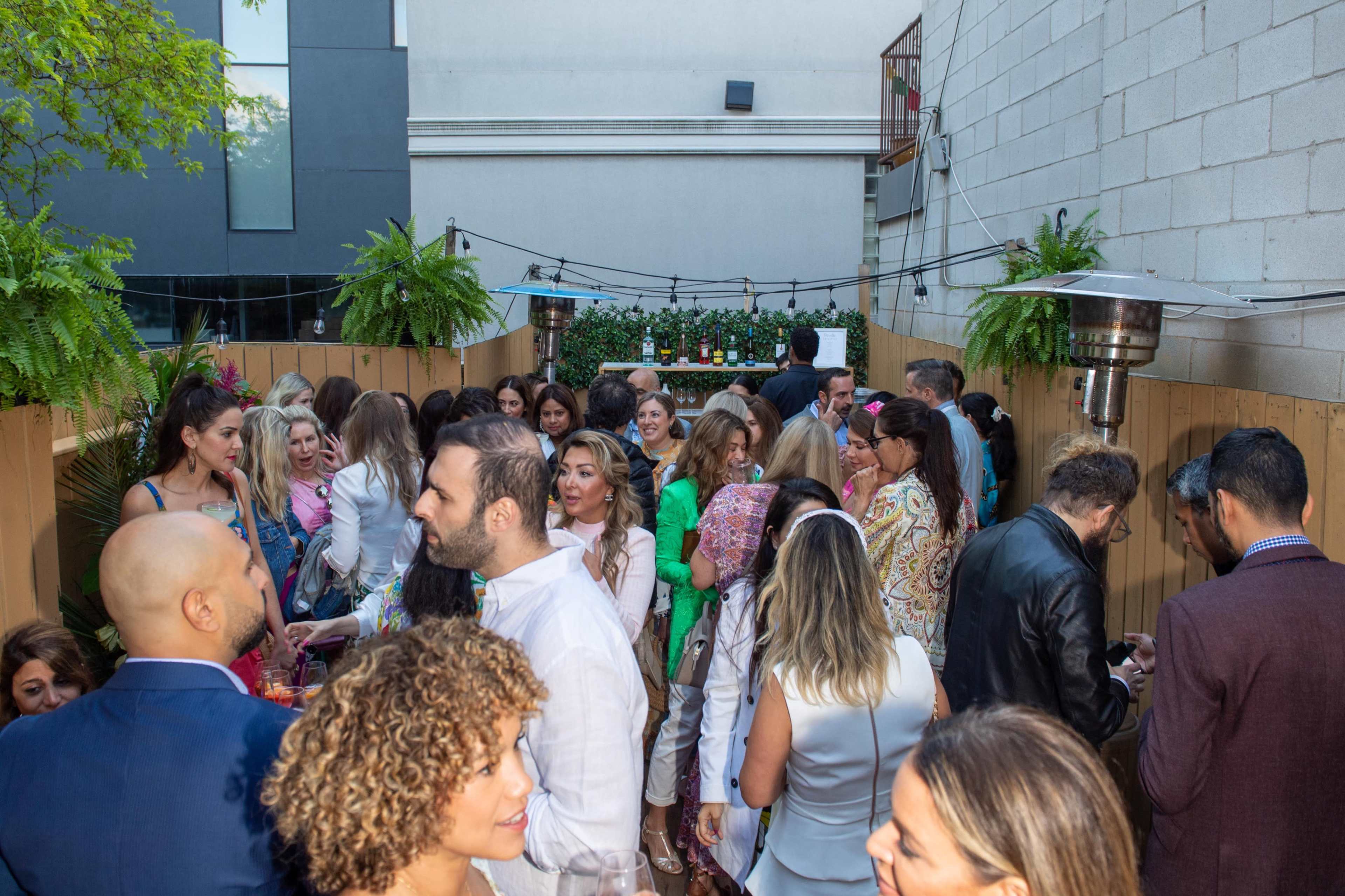 A large group of people socializes at an outdoor gathering surrounded by greenery and wooden fencing.