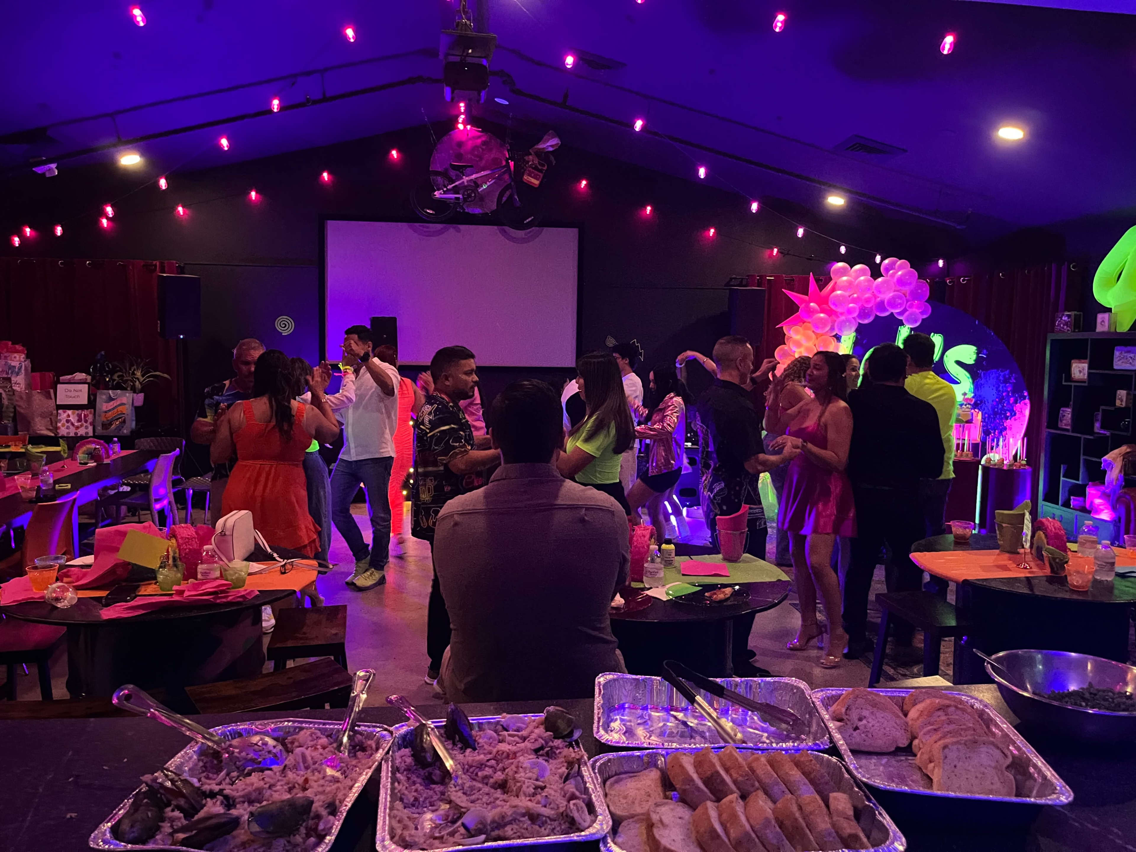 A lively indoor party scene features guests dancing, colorful decorations, and a buffet table with various food items in the foreground.