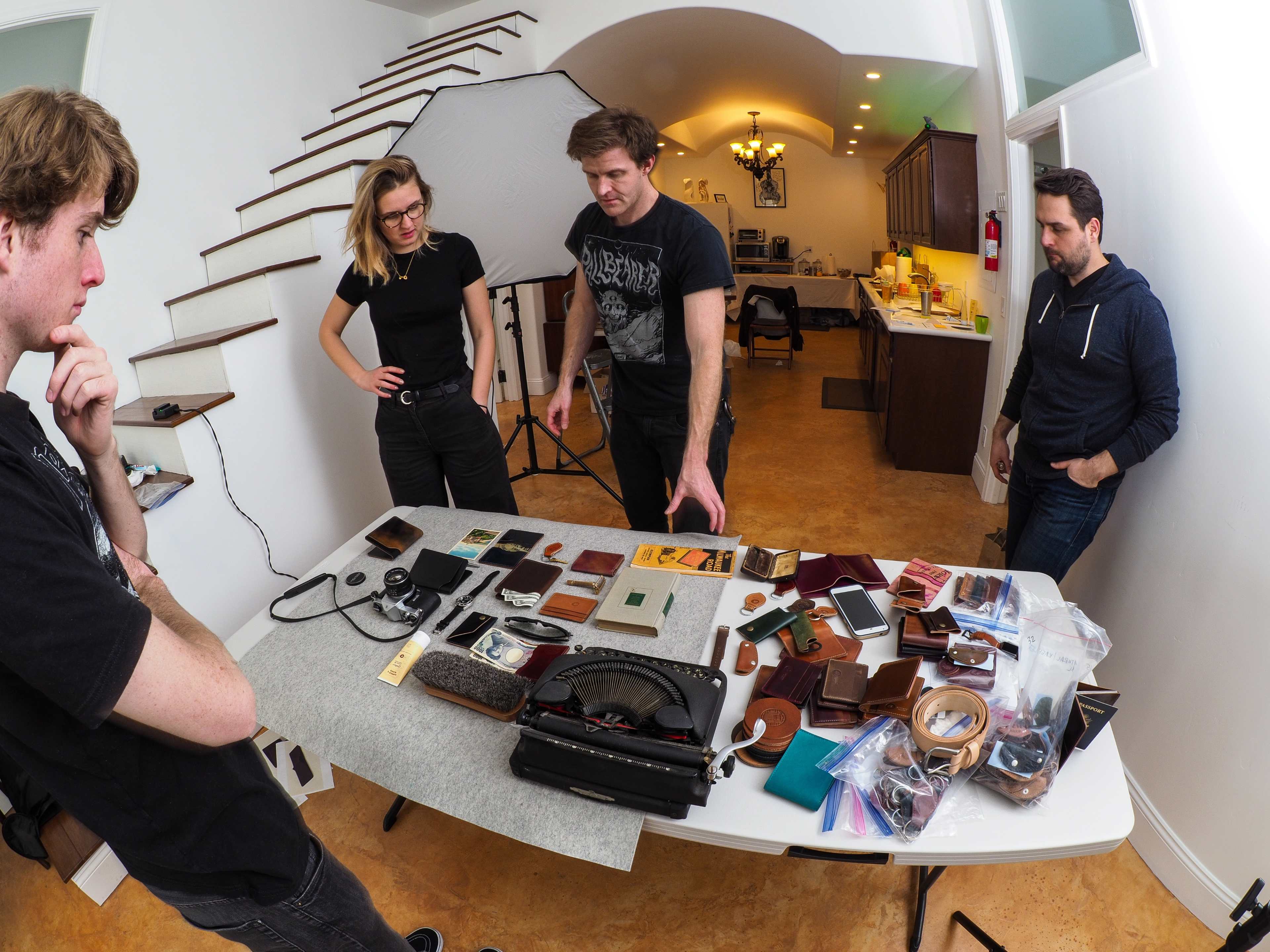 Four individuals examine a table filled with various vintage items and leather goods in a well-lit room.